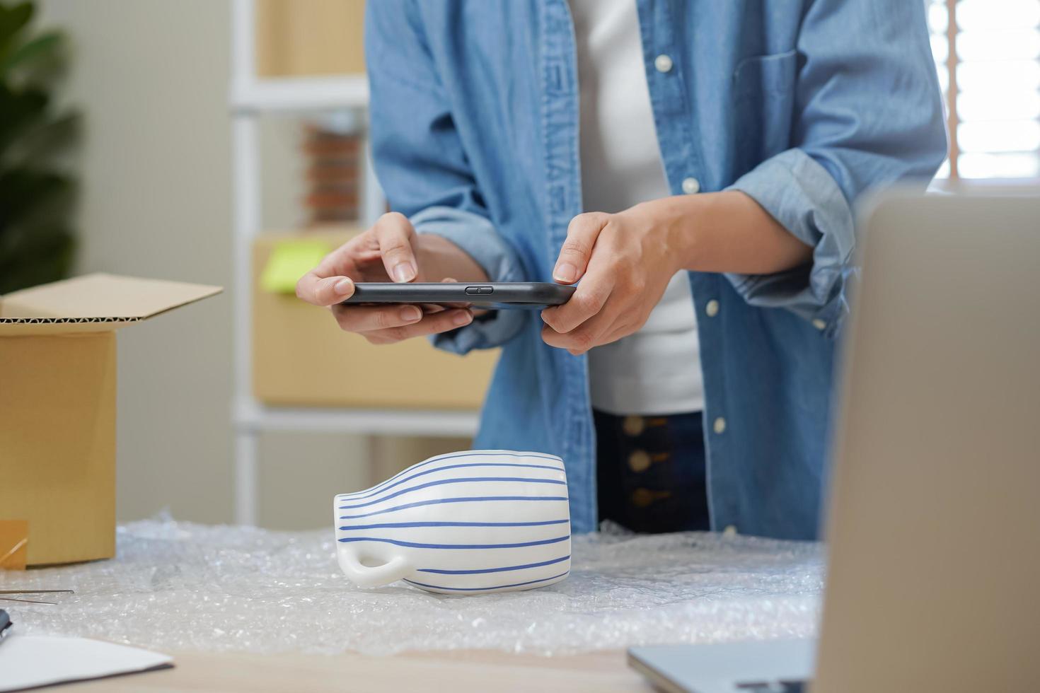 klein bedrijf ondernemer MKB, Aziatisch jong vrouw eigenaar inpakken Product doos, levering, gebruik makend van smartphone nemen een foto, controle pakket voordat sturen bestellen naar klant. handelaar, werk online, e-commerce. foto