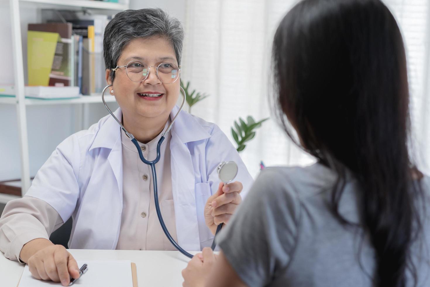 volwassen, senior Aziatisch arts, psychiater, algemeen vrouw dokter overleg plegen bezorgd met jong geduldig, gebruik makend van stethoscoop naar luister, afspraak Bij kliniek, ziekenhuis. Gezondheid zorg, controleren omhoog medisch. foto