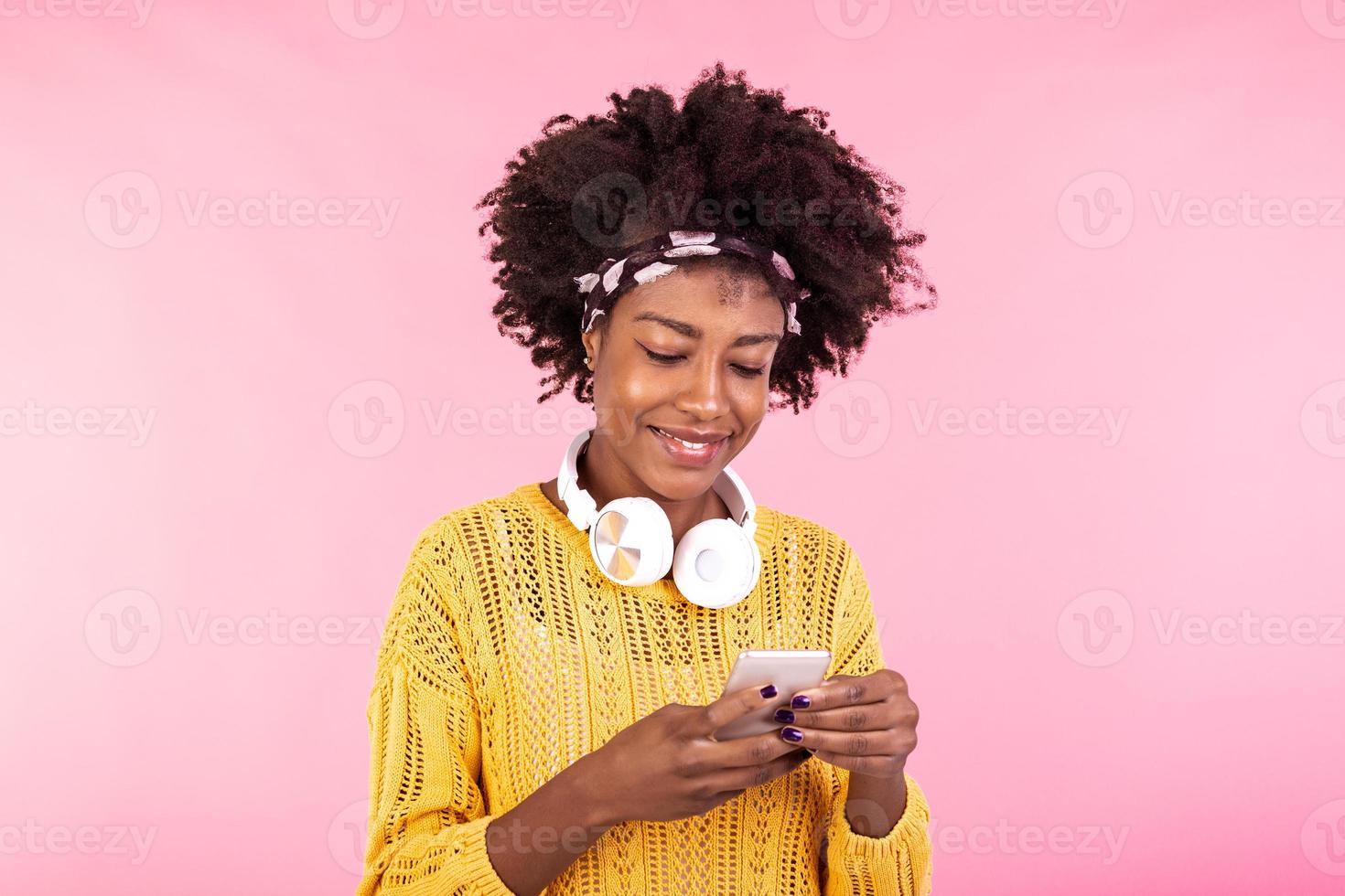 Afrikaanse Amerikaans vrouw vervelend hoofdtelefoons en lachend. jong vrouw met natuurlijk gekruld haar- Holding en op zoek Bij mobiel telefoon. foto