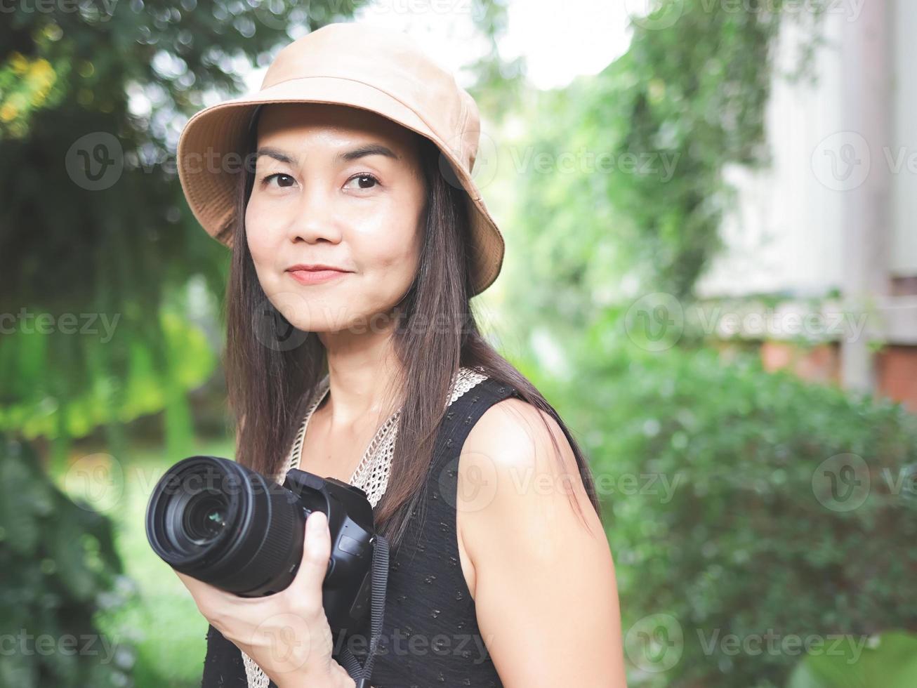 Aziatisch vrouw, vervelend hoed en zwart top mouwloos, staand in de tuin en Holding dslr camera, glimlachen gelukkig. foto