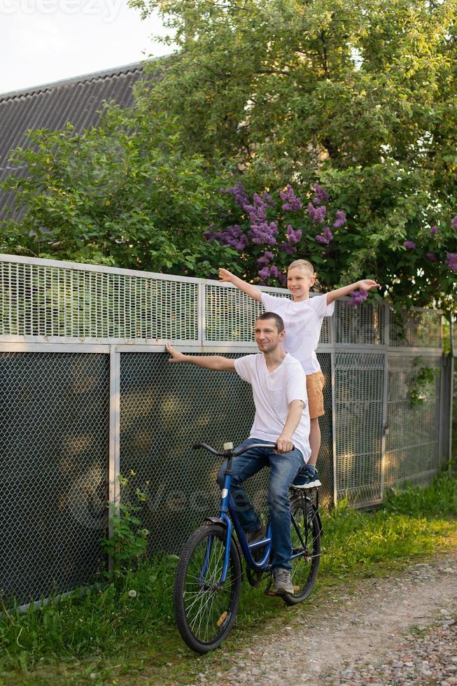 een vader ritten zijn zoon in een wit t-shirt Aan een fiets. de gelukkig zoon verspreiding zijn armen foto