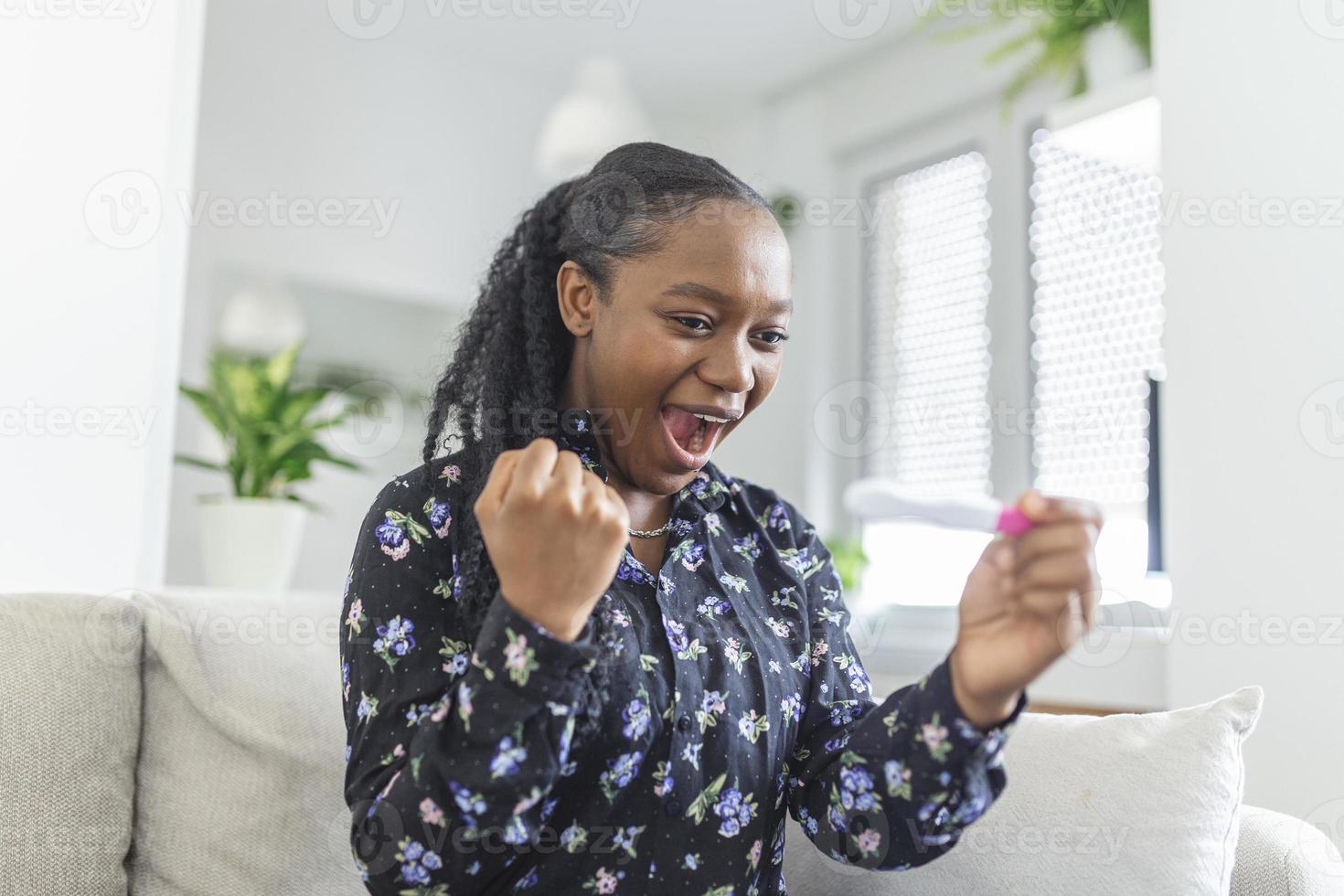 jong vrouw op zoek Bij zwangerschap test in geluk. Tenslotte zwanger. aantrekkelijk zwart Dames op zoek Bij zwangerschap test en glimlachen terwijl zittend Aan de sofa Bij huis foto