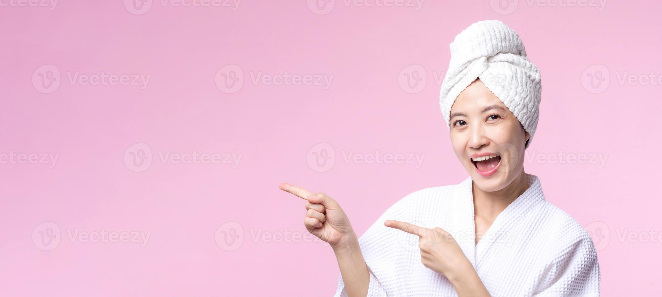 mooi jong Aziatisch vrouw gelukkig glimlach gezicht met hand- of vinger gebaar tekens in badjas en douche handdoek tonen leeg ruimte geïsoleerd Aan roze achtergrond. foto