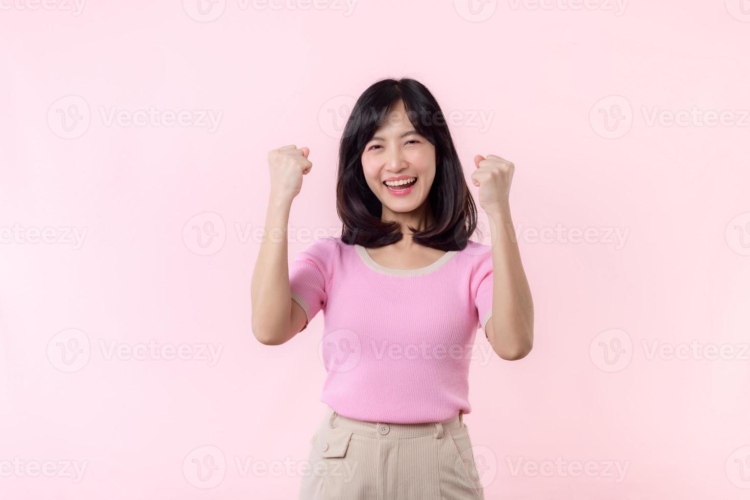 portret mooi jong Aziatisch vrouw gelukkig glimlach met vuist omhoog zege gebaar uitdrukking vrolijk haar succes prestatie tegen roze pastel studio achtergrond. vrouw dag winnaar viering concept. foto