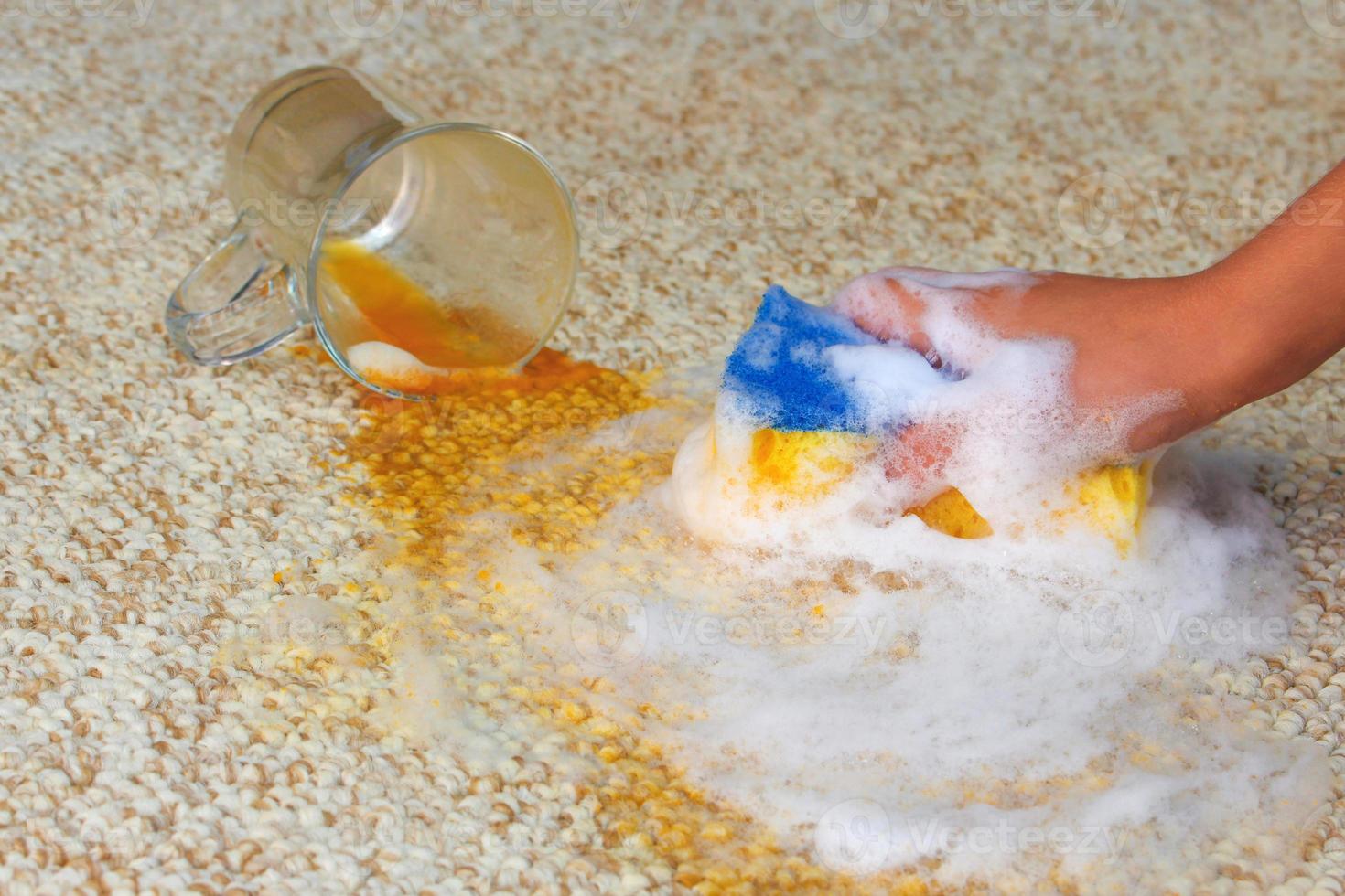 glas van sap viel en gemorst Aan vloer. vrouw hand- reinigt de tapijt met een spons en wasmiddel. foto