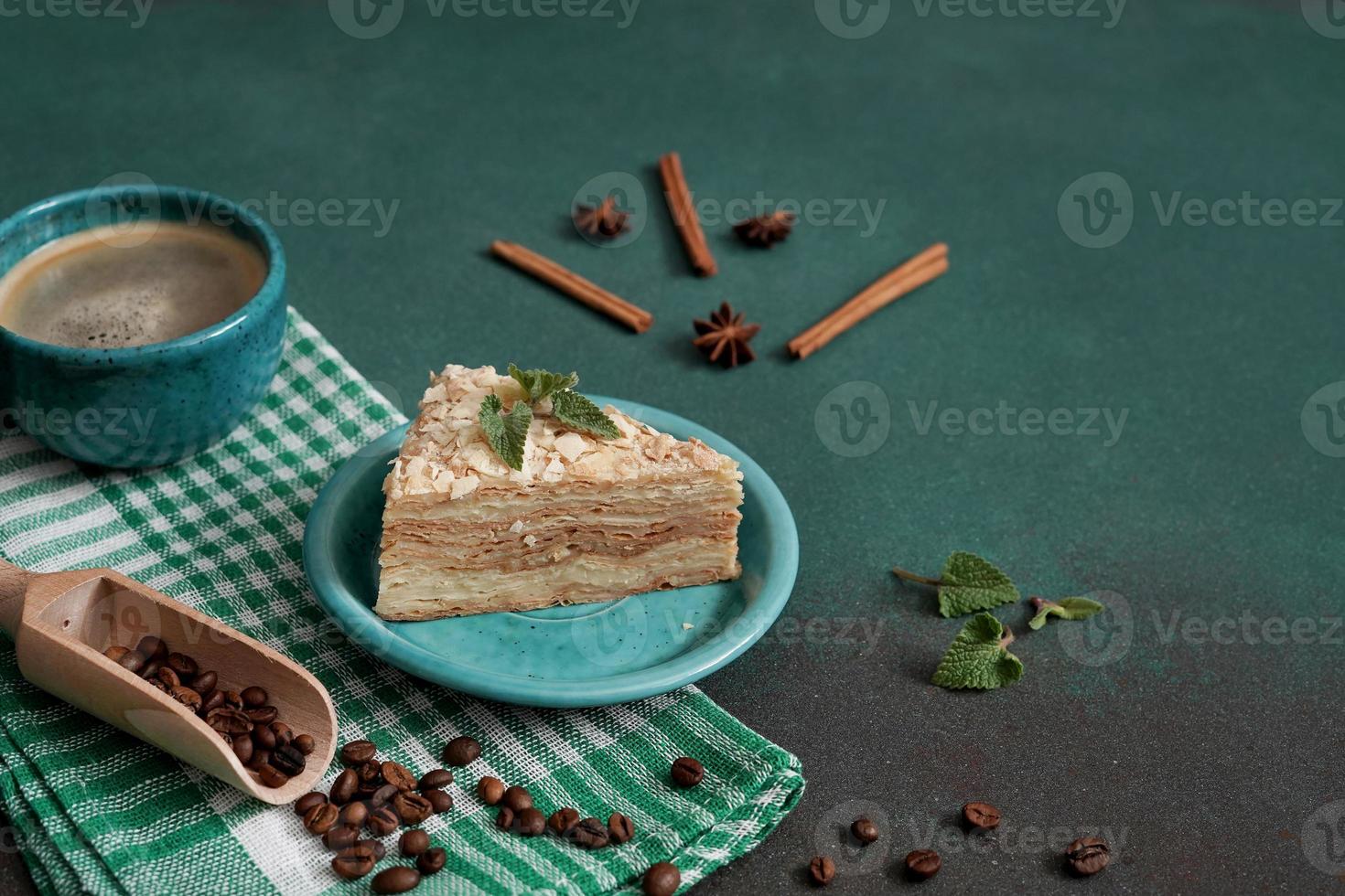 heerlijk Napoleon taart met room Aan een turkoois bord versierd met een takje van munt Aan een groen achtergrond. een kop van heet koffie, kaneel stok, badyan, koffie bonen Aan een groen achtergrond foto