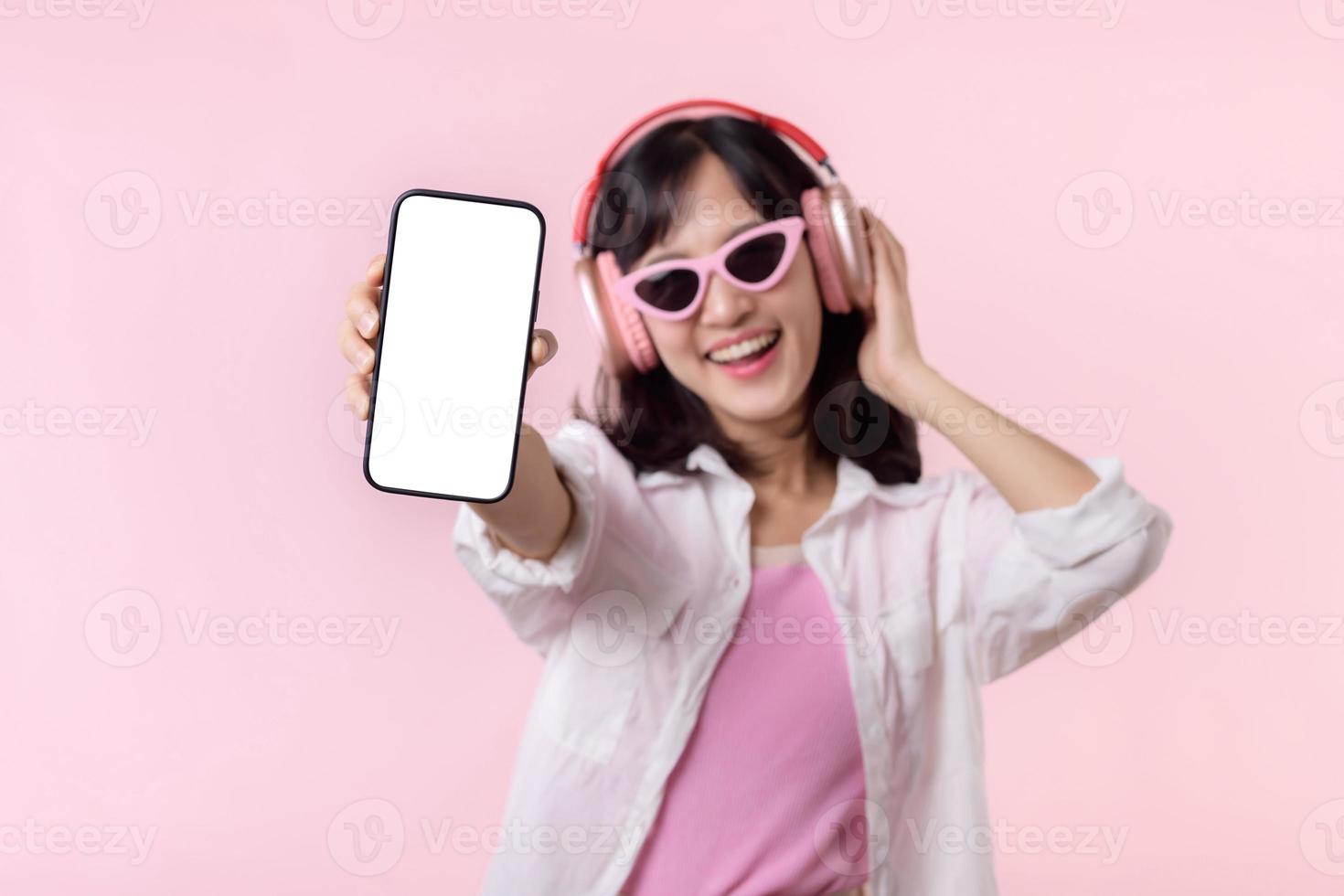 gelukkig vrolijk glimlachen Aziatisch vrouw met draadloze oortelefoons tonen blanco scherm mobiel telefoon of nieuw smartphone muziek- toepassing advertentie mockup geïsoleerd Aan roze studio achtergrond. foto