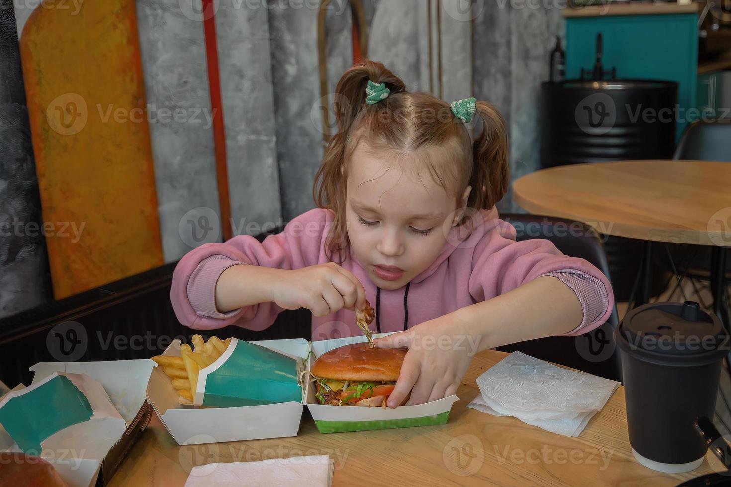 een weinig hongerig meisje met eetlust eet snel voedsel in een cafe Bij een tafel, een hamburger en Frans Patat foto