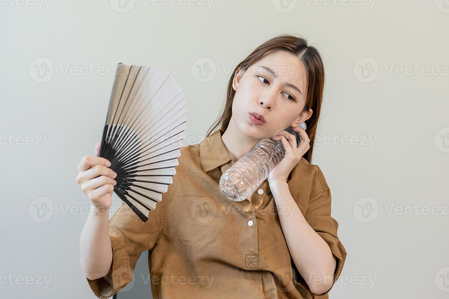 zomer warmte hartinfarct, heet het weer, moe Aziatisch jong vrouw zweterig en dorstig, verfrissend met hand- in blazen, Golf ventilator naar ventilatie, Holding verkoudheid water fles kraan haar lichaam wanneer temperatuur hoog. foto