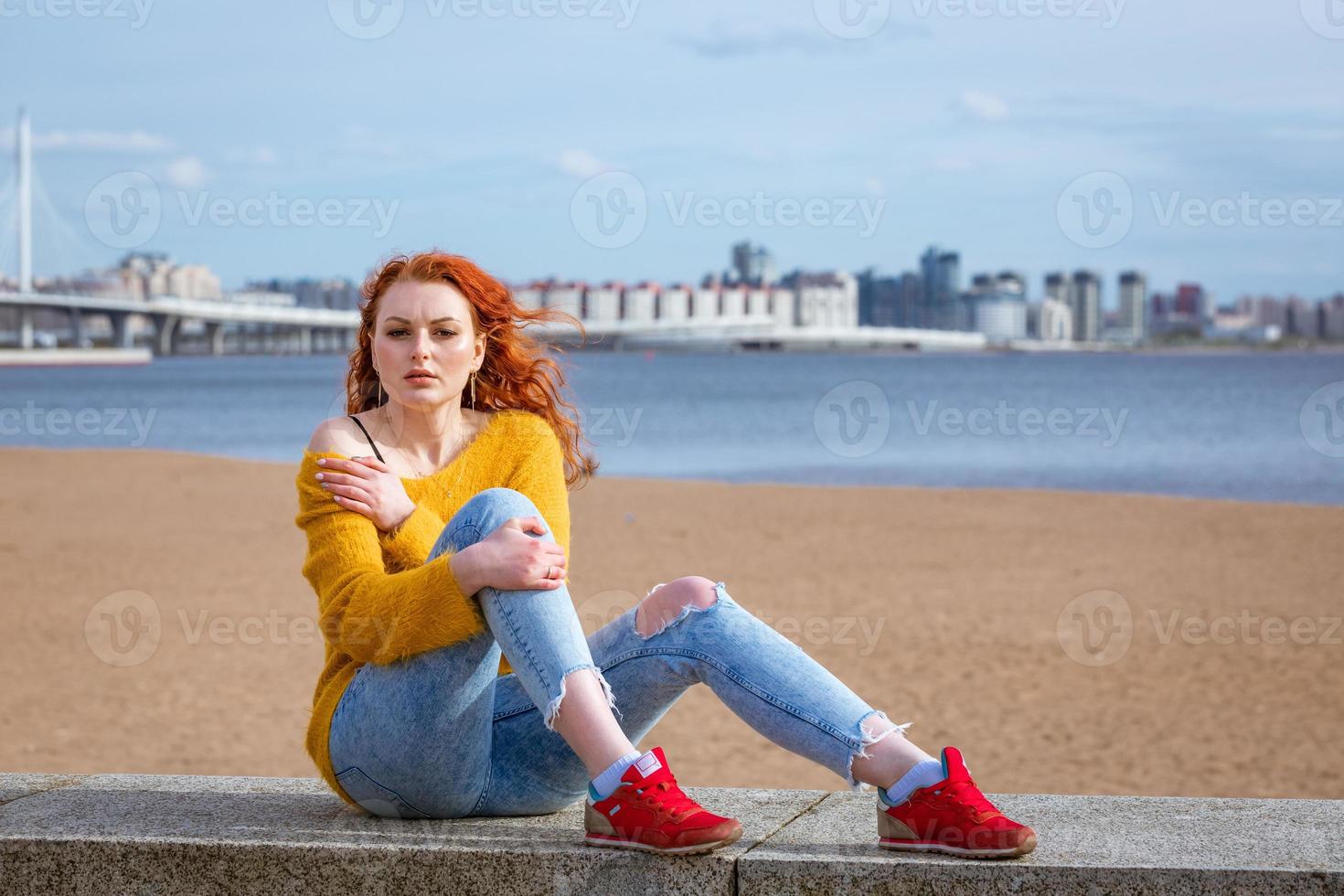 aantrekkelijk jong roodharige vrouw zittend buiten Aan waterkant, vervelend geel foto