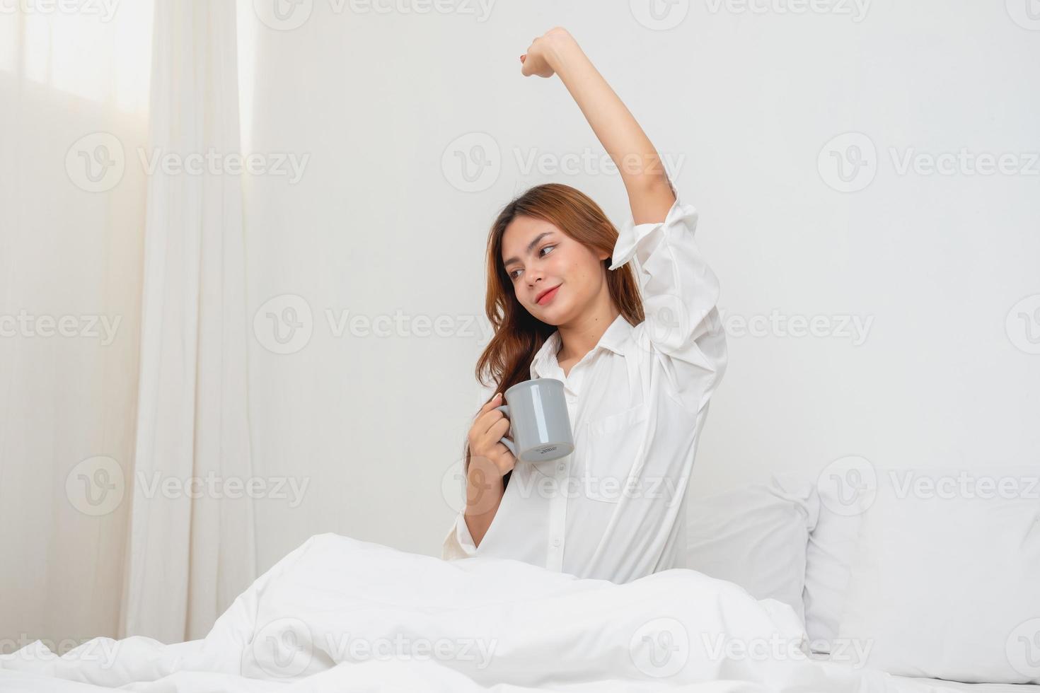 vrouw in wit nachthemd ontwaken omhoog Aan weekend ochtend- resting en ontspannende spelen met laptop mobiel telefoon aan het eten brood en drinken thee in glas binnen wit slaapkamer. ochtend- vakantie concept. foto