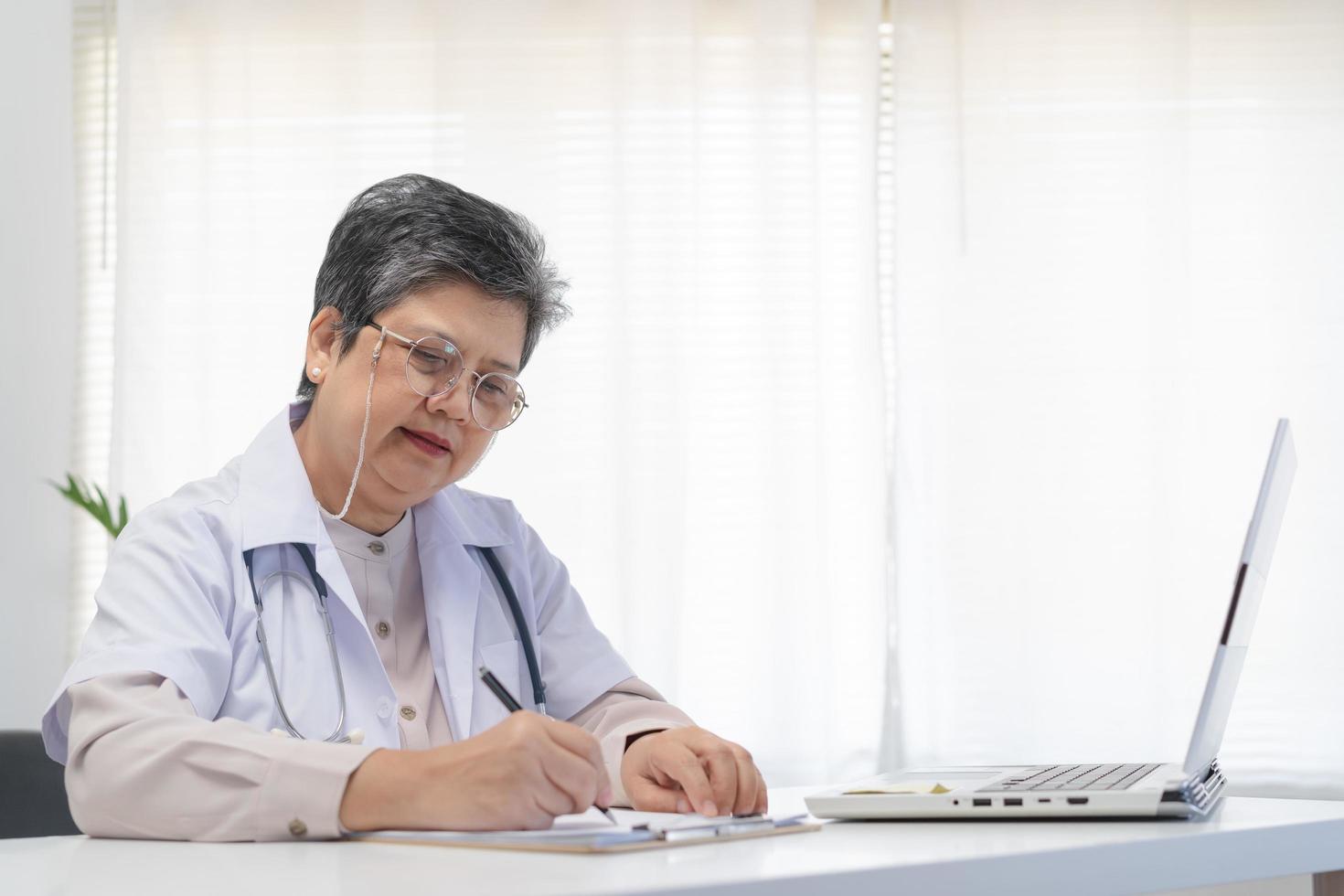 senior Aziatisch arts, algemeen vrouw dokter vervelend bril en wit uniform, volwassen vrouw met stethoscoop, werken Aan laptop computer, zittend in medisch kantoor. gezondheidszorg en medisch. foto