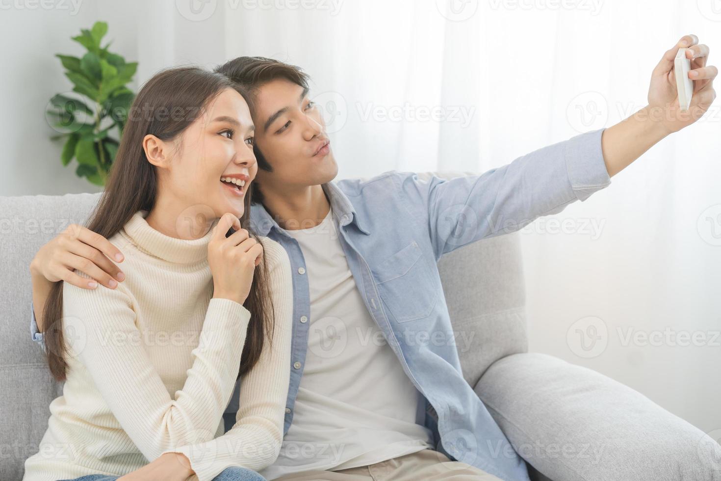 gelukkig paar liefde Bij huis, twee Aziatisch jong uitgeven tijd, bonding naar elk andere romantisch Aan sofa in leven kamer terwijl Mens omhelzing vrouw gebruik makend van smartphone, mobiel telefoon naar selfie nemen een foto samen.