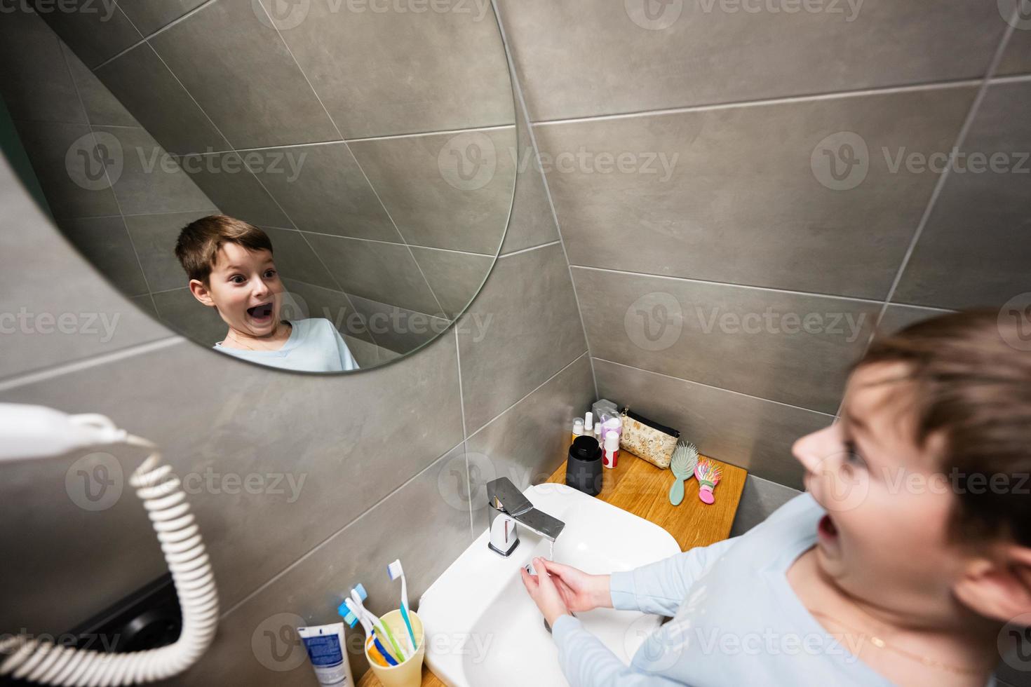 jongen wast met grappig gezicht in spiegel Bij badkamer. foto