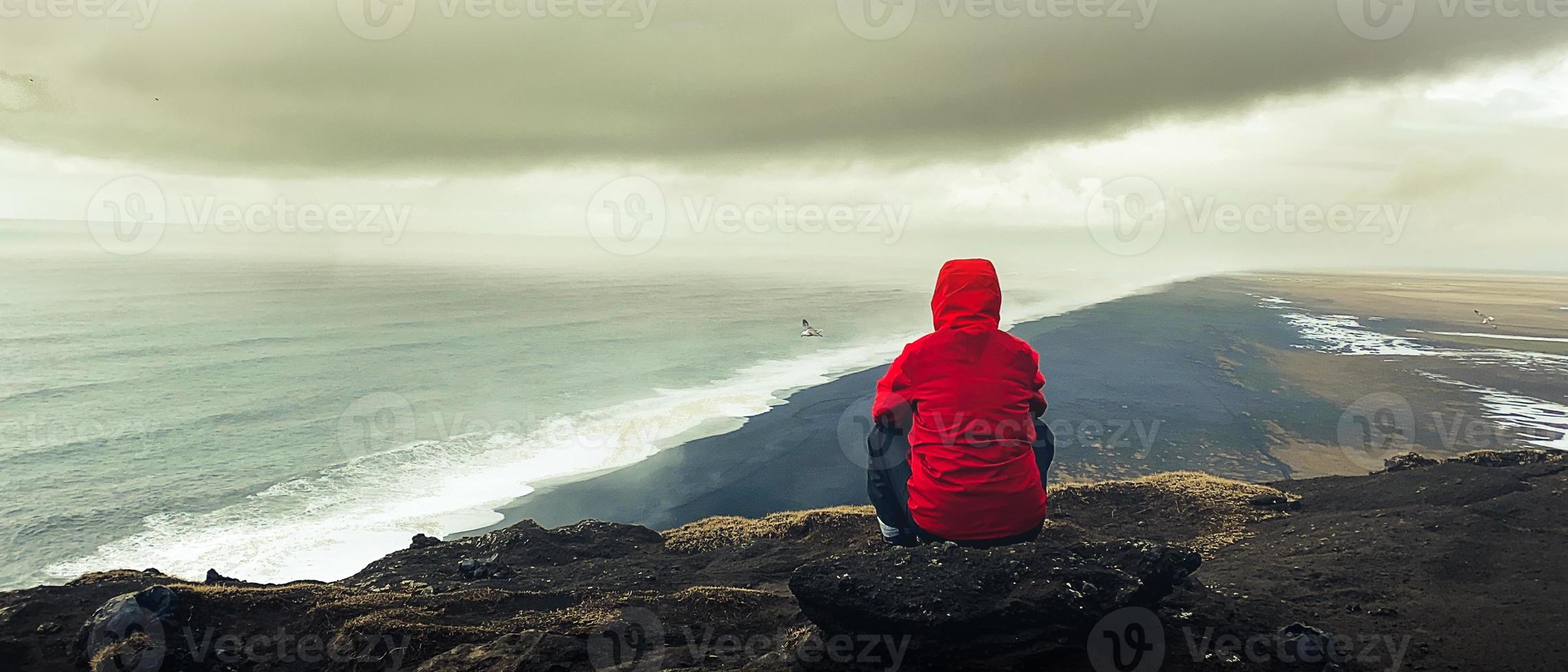 Mens zitten Aan rots attent kijken Bij atlantic oceaan golven. beroemd iconisch klif gezichtspunt over- reynisfjara zwart zand strand. persoon looks voor richting en doel Aan reist foto