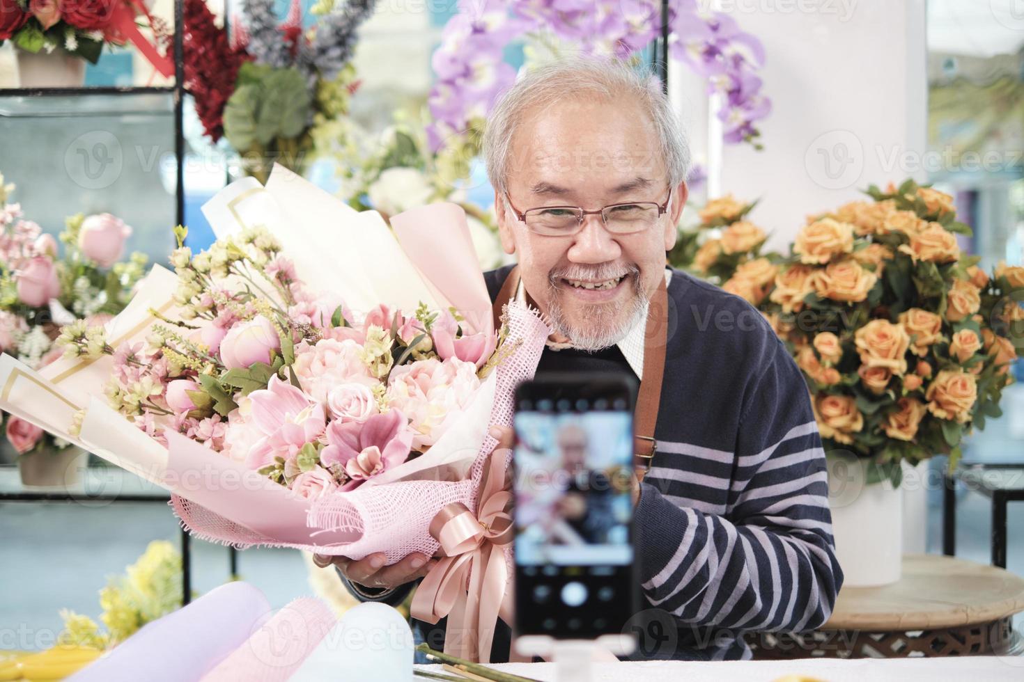 e-commerce bedrijf, een senior mannetje bloemist arbeiders demonstreren en tonen bloemen regelingen via online leven streaming met smartphone toepassing in een helder bloem winkel, een mooi bloesems op te slaan. foto