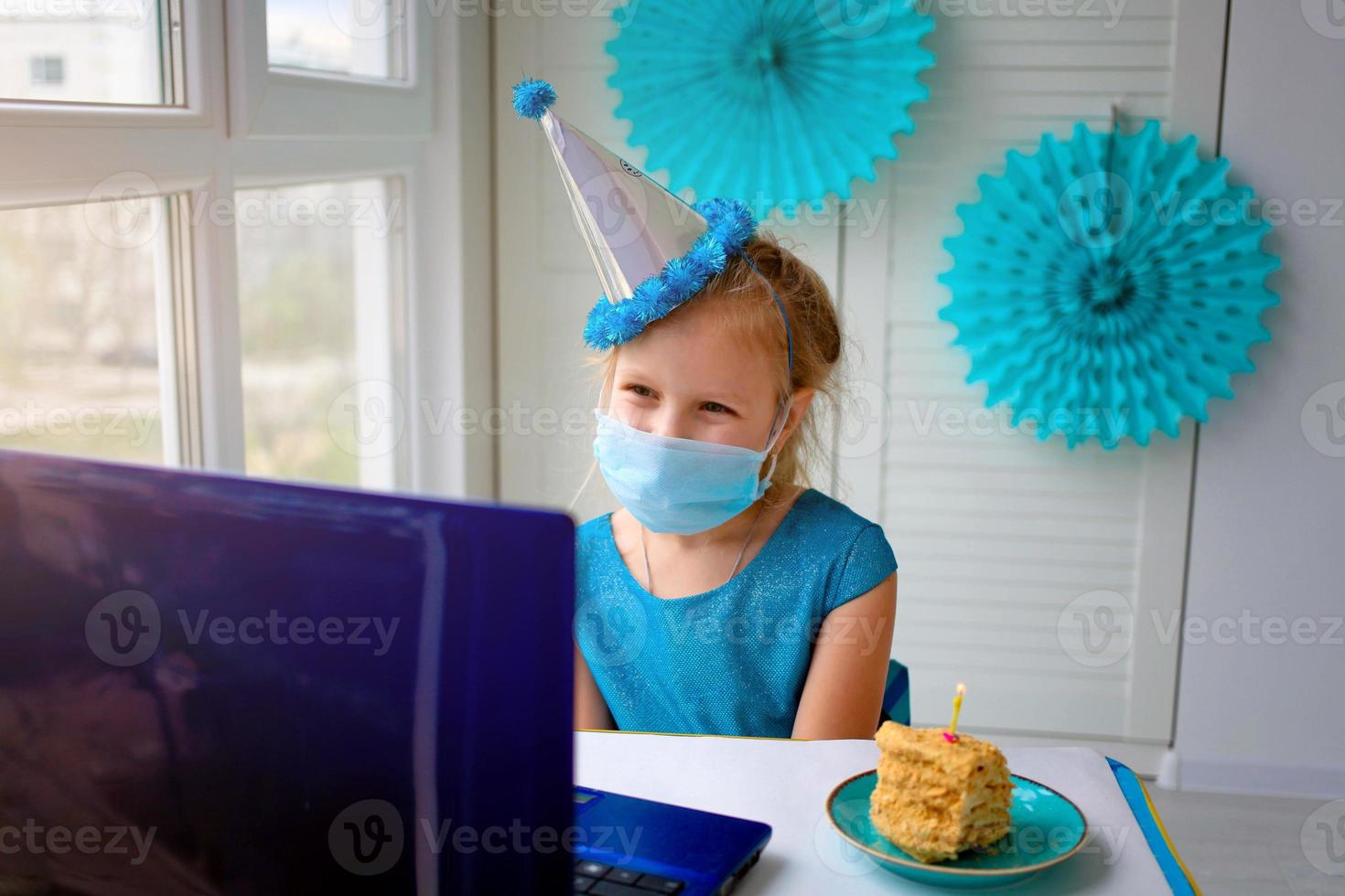 een gelukkig weinig meisje, vervelend een medisch masker in voorkant van haar laptop viert haar verjaardag via de internet in quarantaine tijd, zelfisolatie, online verjaardag. foto