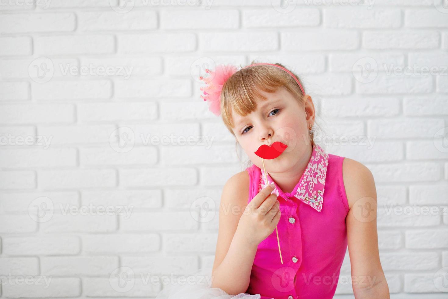 mooi weinig meisje, vervelend Aan roze jurk, poseren met rood papier lippen, met een kroon Aan zijn hoofd Aan wit achtergrond, taille omhoog. foto