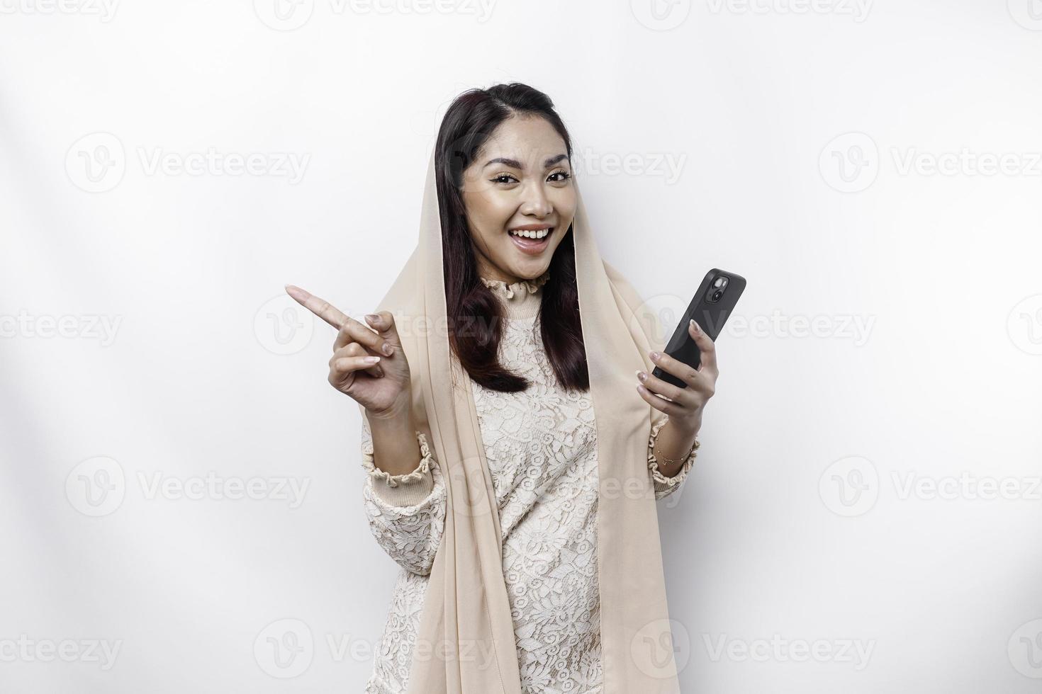 opgewonden Aziatisch vrouw vervelend hijab richten Bij de kopiëren ruimte naast haar terwijl Holding haar telefoon, geïsoleerd door wit achtergrond foto