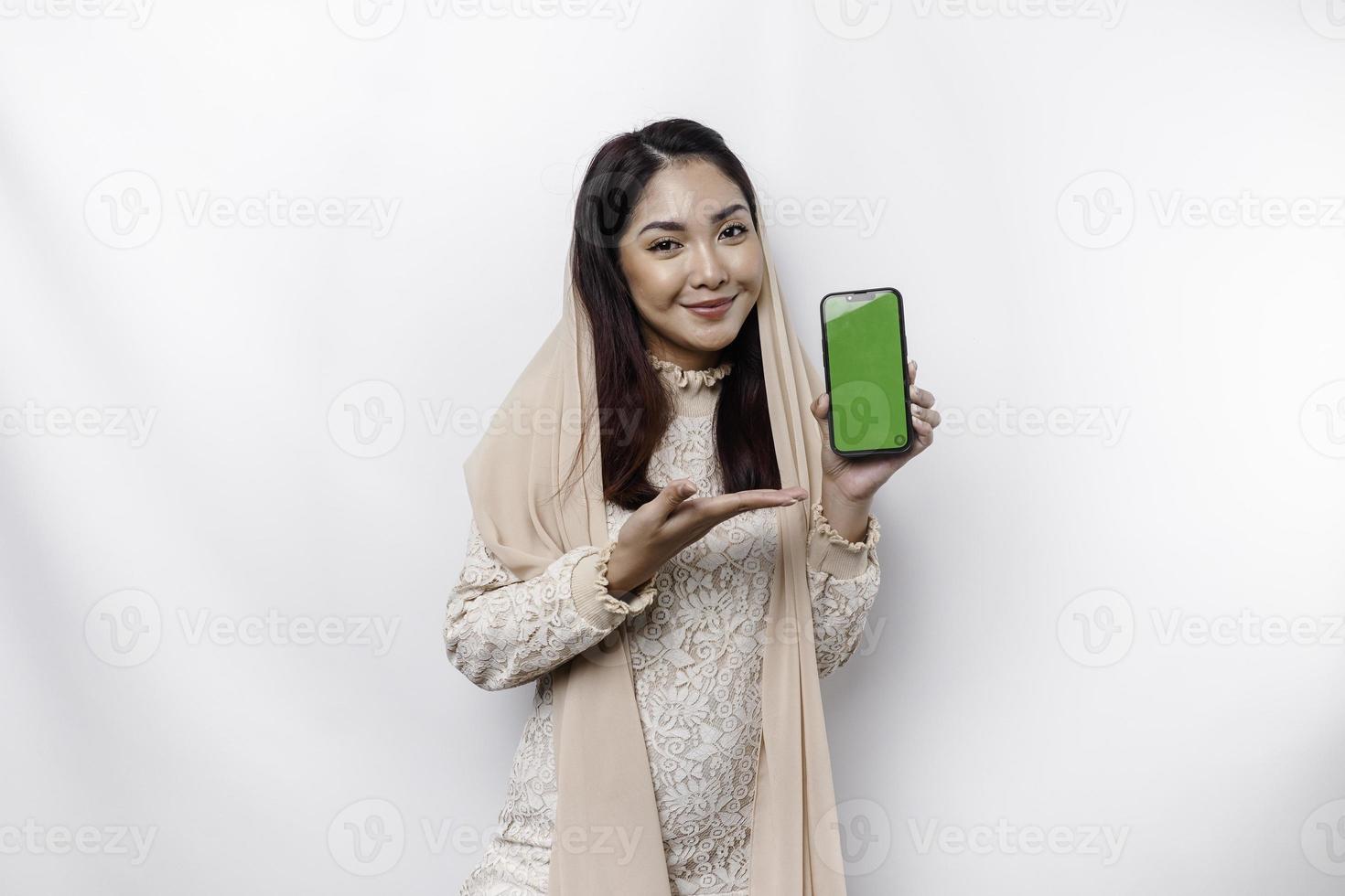 een portret van een gelukkig Aziatisch moslim vrouw vervelend een hoofddoek, tonen haar telefoon scherm, geïsoleerd door wit achtergrond foto