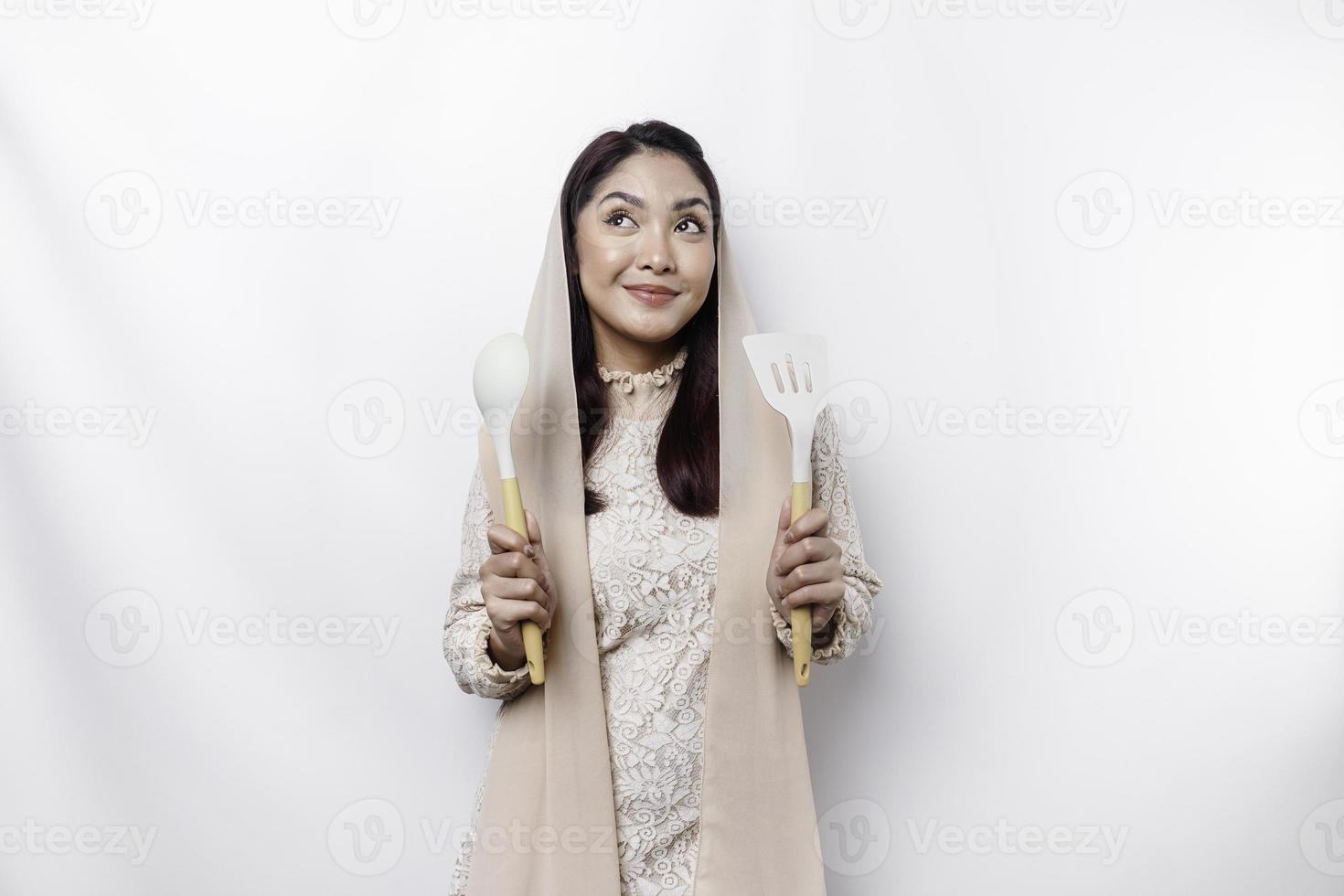 opgewonden Aziatisch moslim vrouw vervelend een hoofddoek, Holding Koken ware en lachend, geïsoleerd door wit achtergrond foto