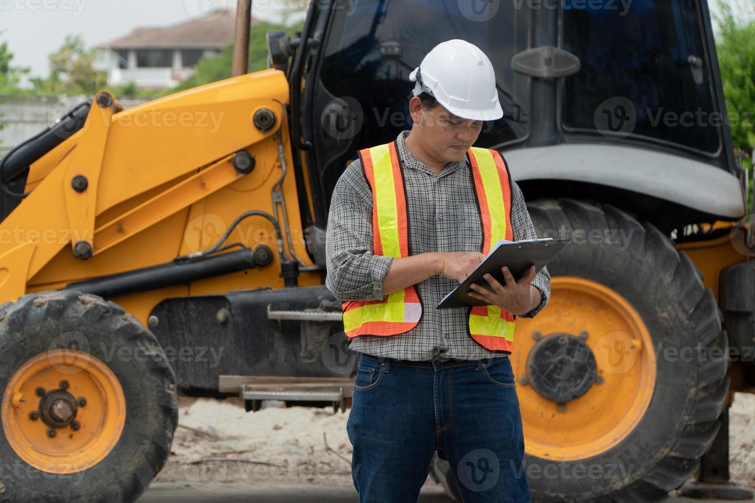 bouwkunde vervelend een wit veiligheid helm staand in voorkant van de backhoe op zoek Bij huis bouw werk en gebruik de tablet naar controleren de blauwdruk met bouw foto