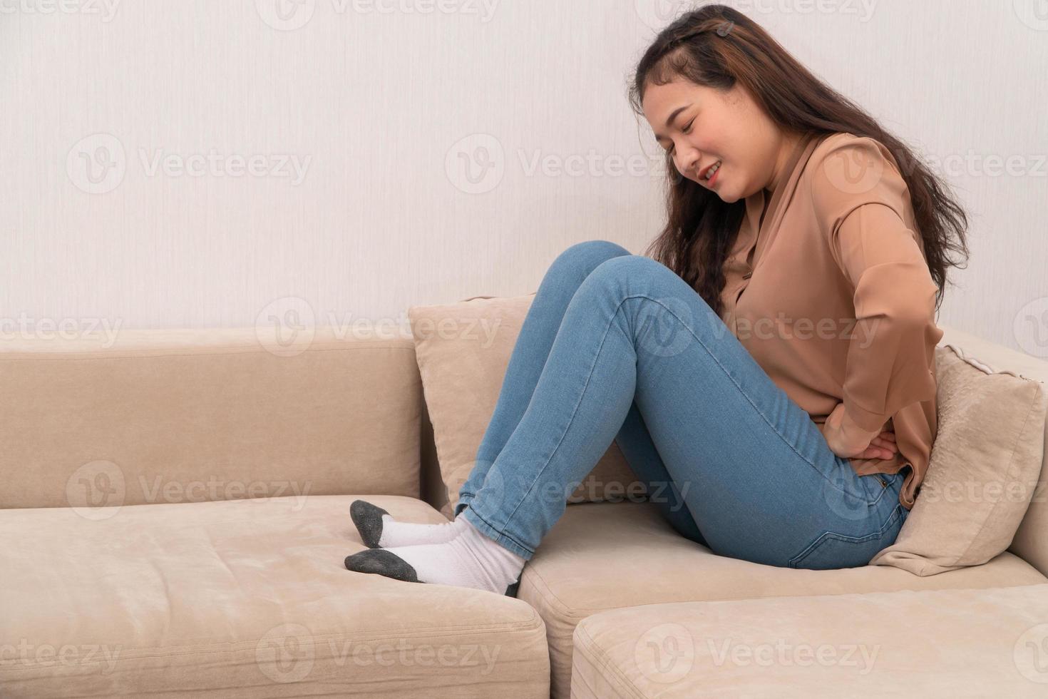 ongelukkige aziatische vrouw die op de bank zit en maagpijn vasthoudt. buikpijn die het gevolg is van menstruatie, diarree of indigestie. ziekte en zorgconcept foto