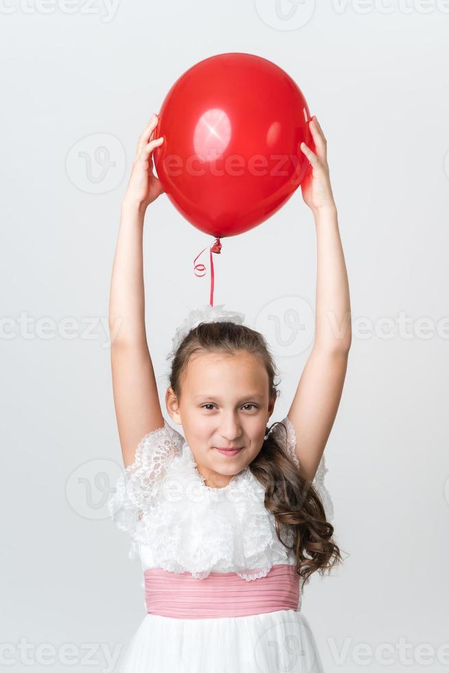 speels meisje in wit jurk houdt rood ballon bovenstaand haar hoofd met beide handen en op zoek Bij camera foto