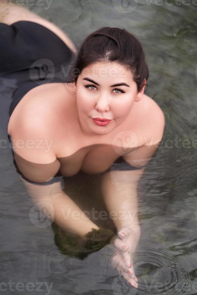 hoog hoek schot van plus grootte vrouw met groot borst in zwart zwempak aan het liegen in buitenshuis zwembad Bij spa foto
