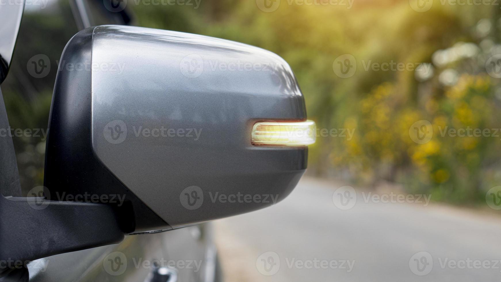 spiegel vleugel van grijs met beurt licht signaal. wazig van asfalt weg en bomen met geel bloem voor achtergrond. foto