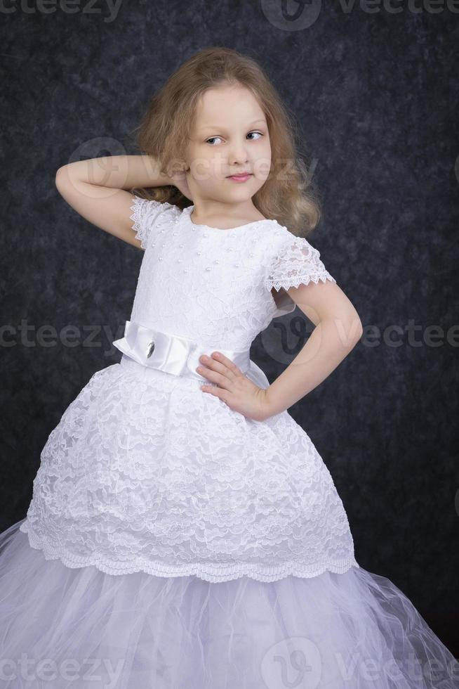 schattig weinig blond meisje in een mooi wit jurk Aan een donker achtergrond. zes jaar oud mooi meisje foto