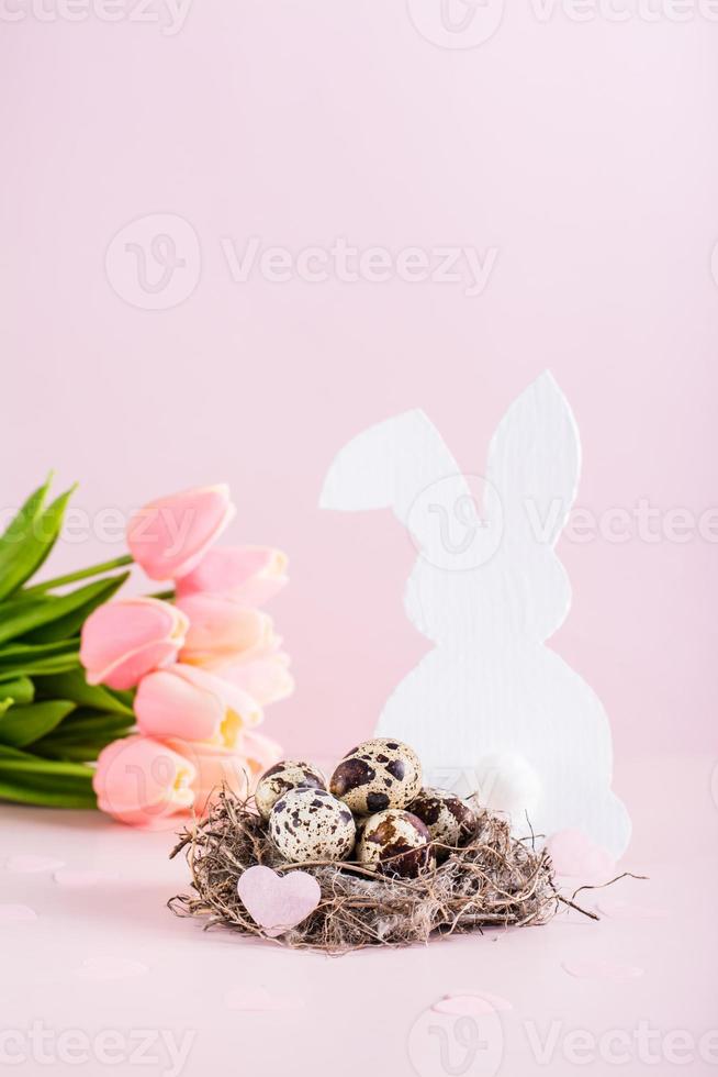 gelukkig Pasen kwartel eieren in een nest, wit konijn en tulpen Aan een roze achtergrond verticaal visie foto