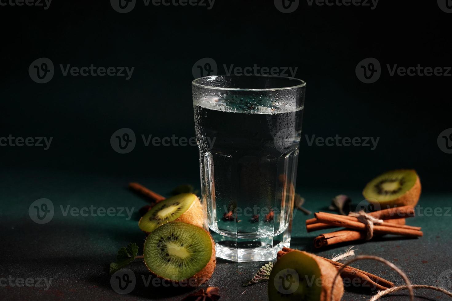 een glas van verkoudheid water Aan een groen achtergrond. versierd met plak kiwi, kaneel stokken, badyan, munt bladeren. kopiëren ruimte foto