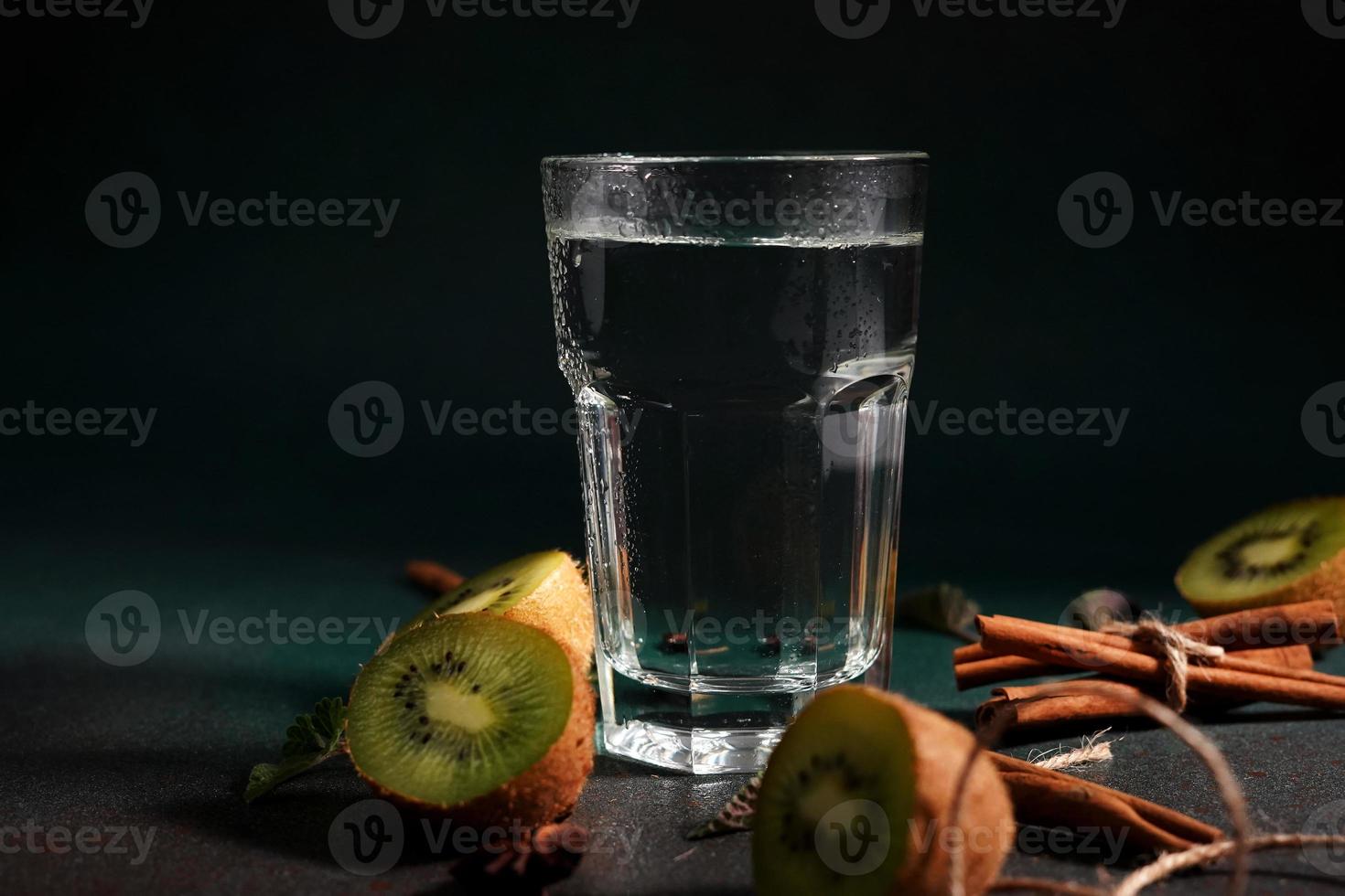 een glas van verkoudheid water Aan een groen achtergrond. versierd met plak kiwi, kaneel stokken, badyan, munt bladeren. kopiëren ruimte foto