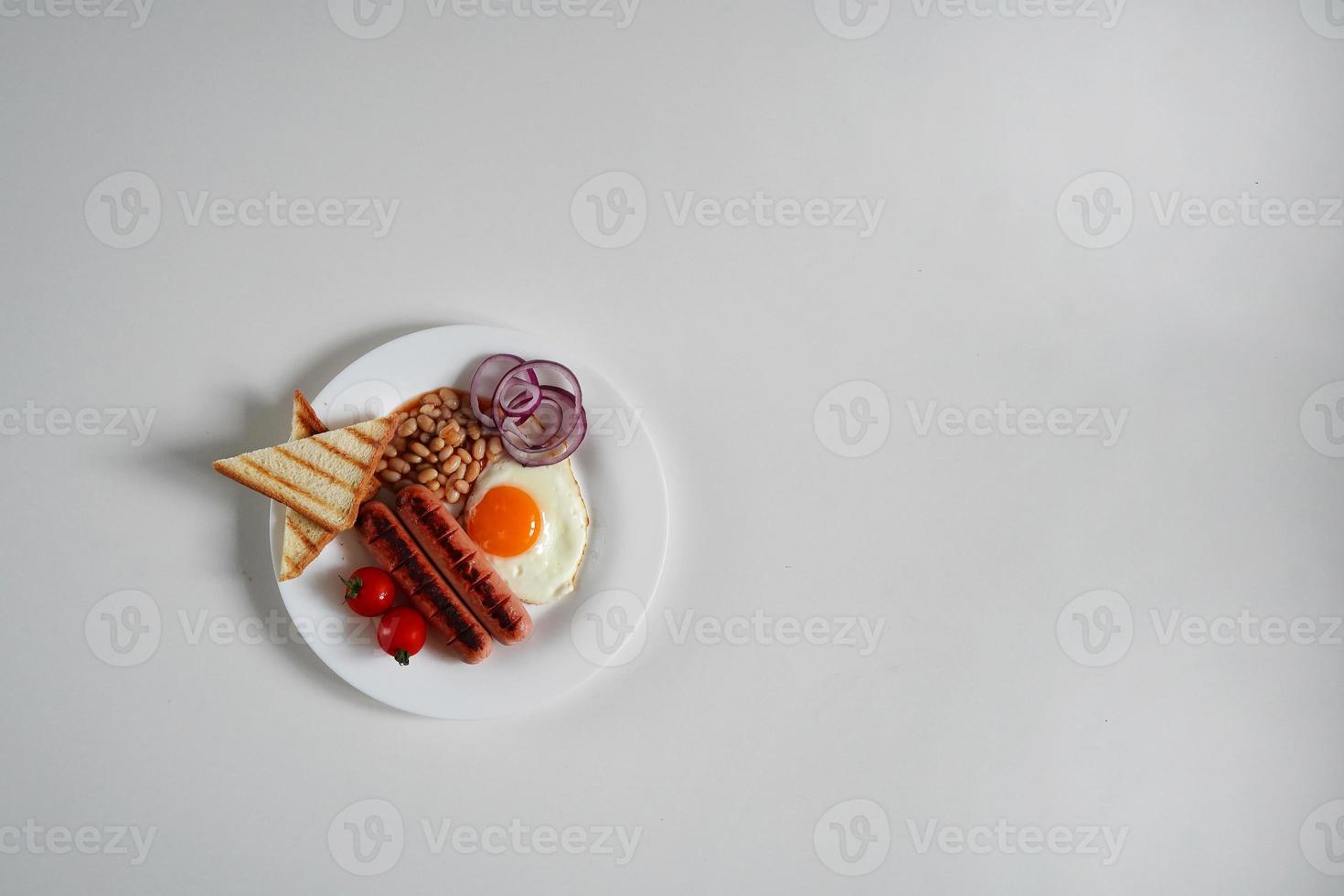 top visie Engels ontbijt. gebakken ei, twee gebakken worstjes, gegrild brood geroosterd brood, ingeblikt bonen, blauw uien en kers tomaten Aan een wit achtergrond foto