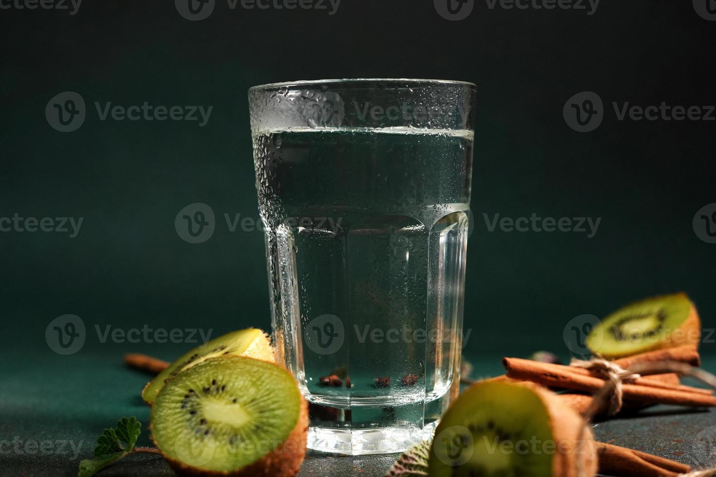 een glas van verkoudheid water Aan een groen achtergrond. versierd met plak kiwi, kaneel stokken, badyan, munt bladeren. kopiëren ruimte foto