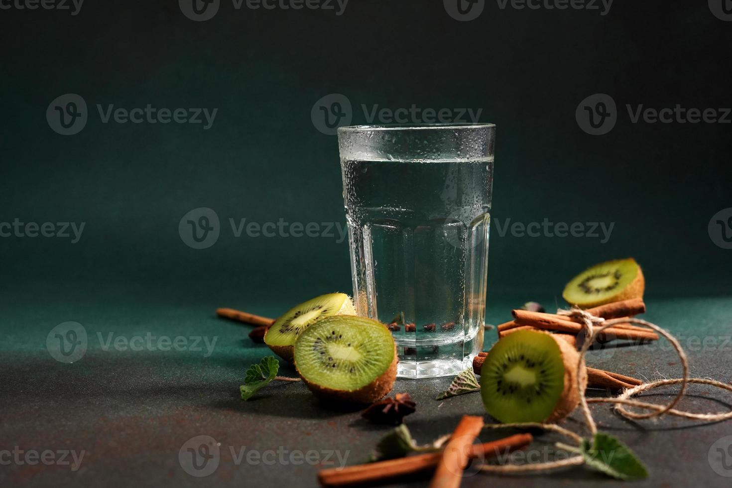 een glas van verkoudheid water Aan een groen achtergrond. versierd met plak kiwi, kaneel stokken, badyan, munt bladeren. kopiëren ruimte foto