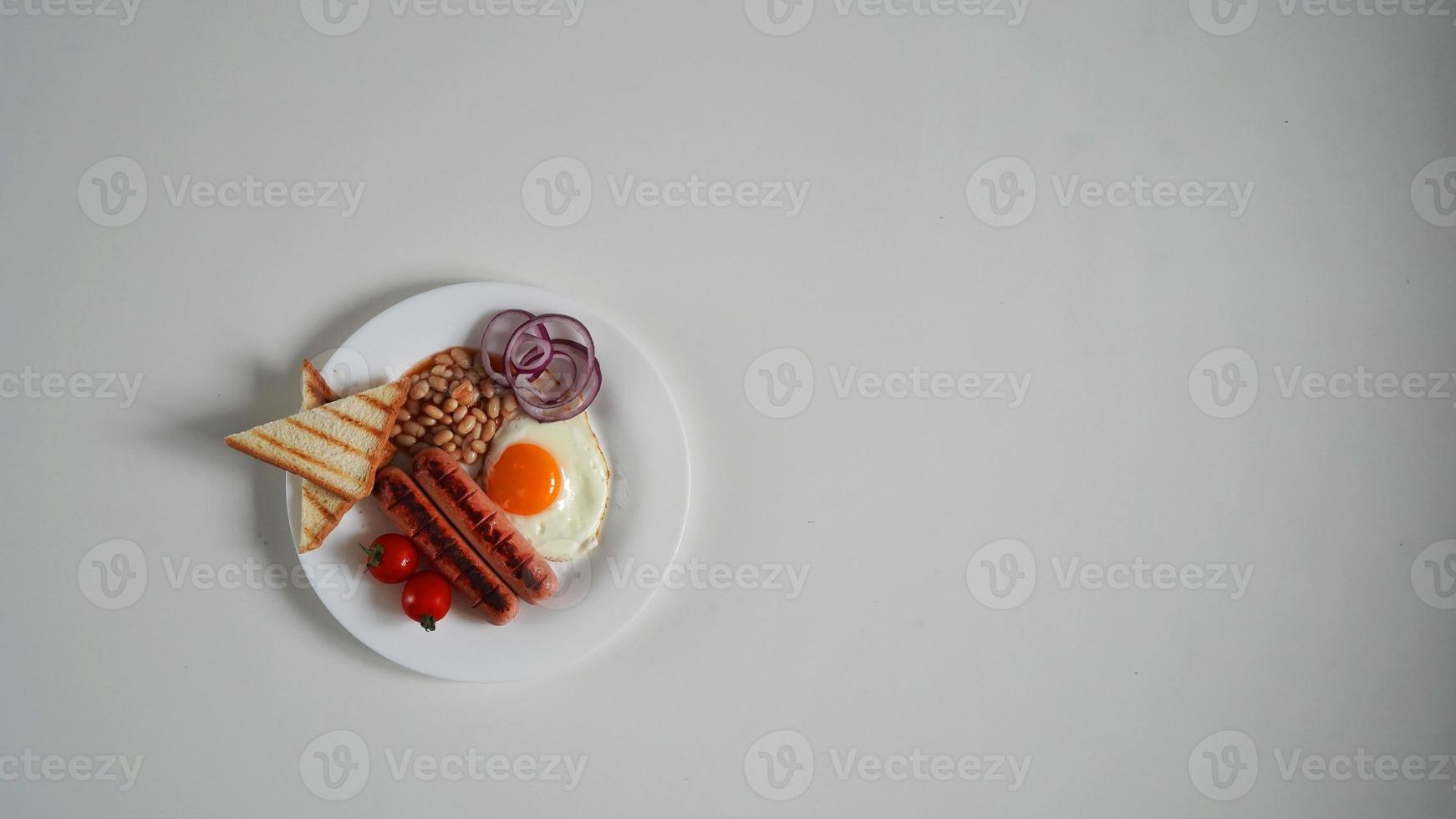 top visie Engels ontbijt. gebakken ei, twee gebakken worstjes, gegrild brood geroosterd brood, ingeblikt bonen, blauw uien en kers tomaten Aan een wit achtergrond foto