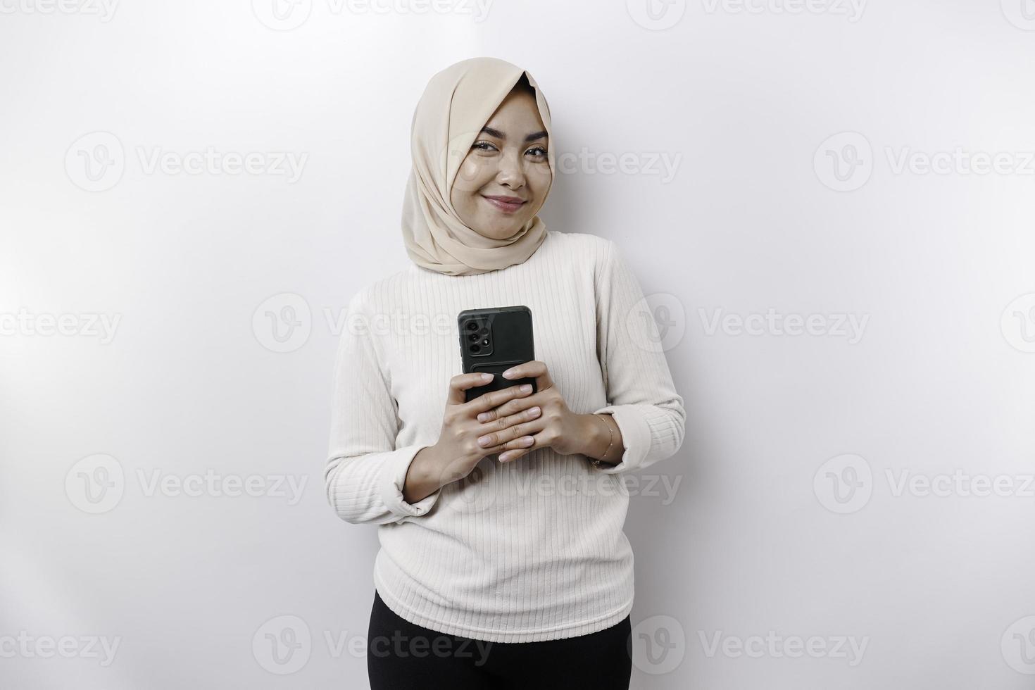 een gelukkig Aziatisch moslim vrouw vervelend een hoofddoek, Holding haar telefoon, geïsoleerd door wit achtergrond foto