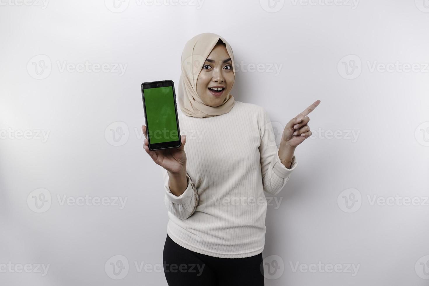 opgewonden Aziatisch moslim vrouw vervelend hoofddoek richten Bij de kopiëren ruimte naast haar terwijl tonen haar telefoon, geïsoleerd door wit achtergrond foto