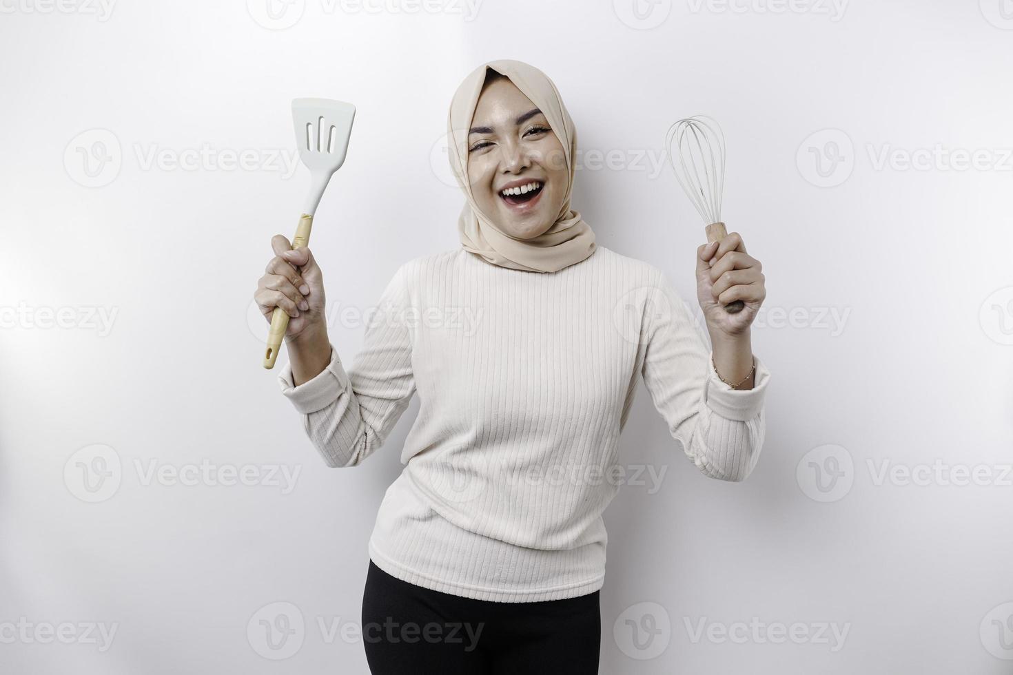 opgewonden Aziatisch moslim vrouw vervelend een hoofddoek, Holding Koken ware en lachend, geïsoleerd door wit achtergrond foto