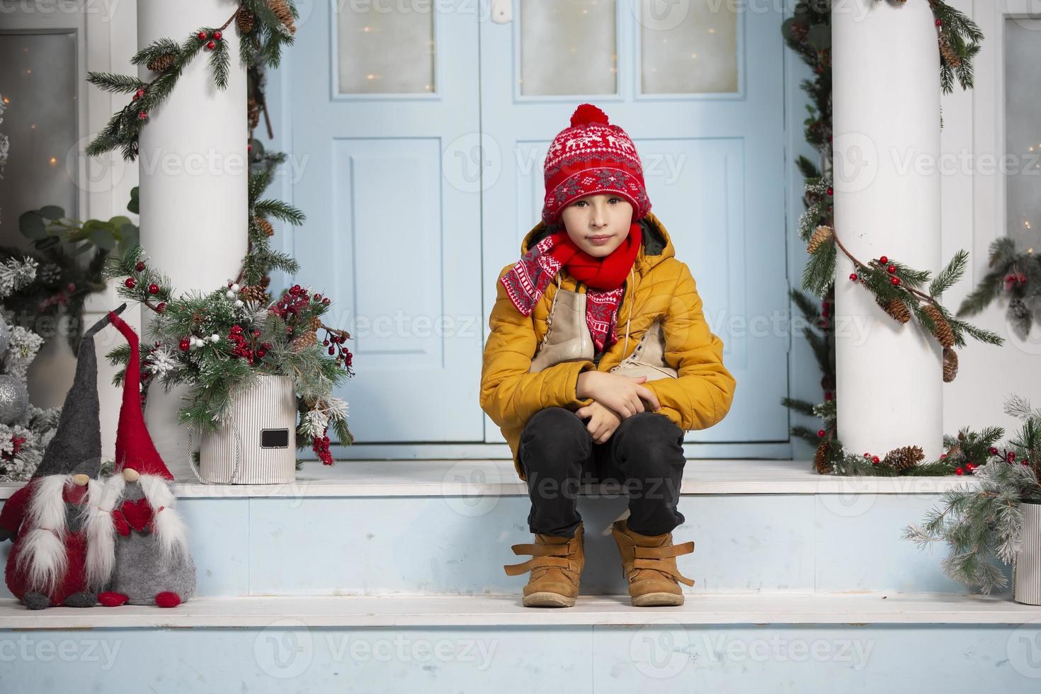 knap weinig jongen in kerstmis. grappig kind houdt oud skates en zit Aan de veranda van een huis versierd met Kerstmis boom decoraties foto