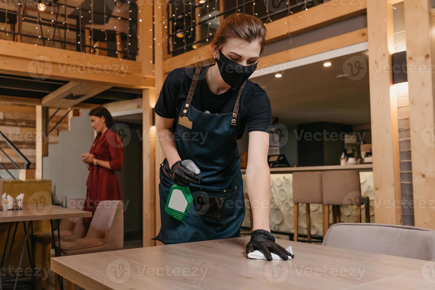 een vriendelijke serveerster die een zwart medisch gezichtsmasker en medische wegwerphandschoenen draagt, houdt een fles met ontsmettingsmiddel vast en maakt tafels schoon met een doek in een restaurant foto