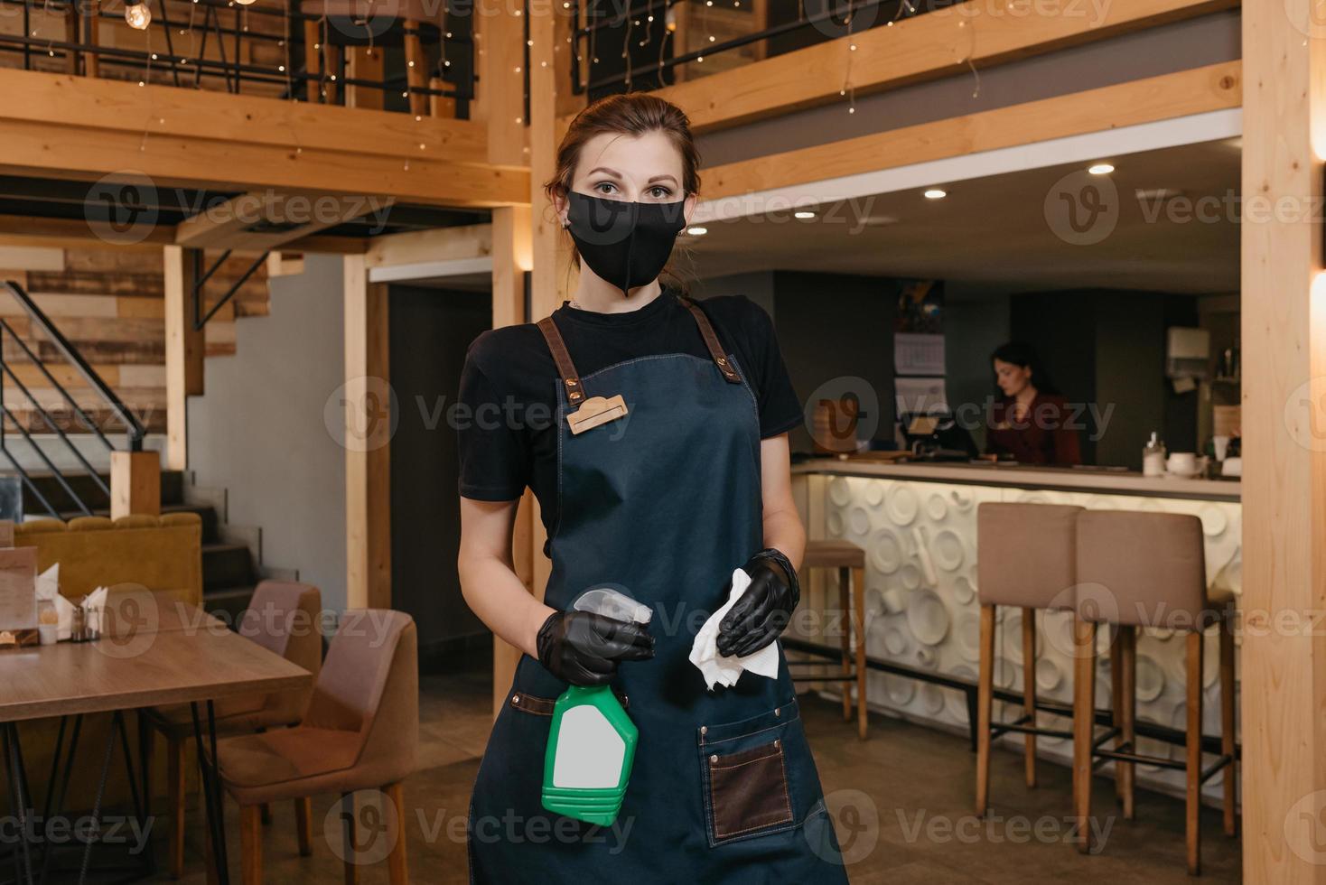 een serveerster met een medisch gezichtsmasker houdt een fles met ontsmettingsmiddel en een doek vast foto