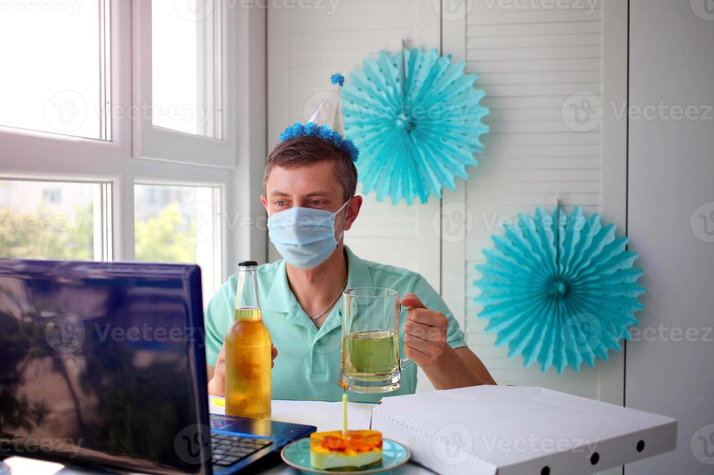 een Mens in een medisch masker zit in voorkant van een laptop, viert een verjaardag met vrienden over- de internet en houdt een glas van alcohol in zijn handen. foto