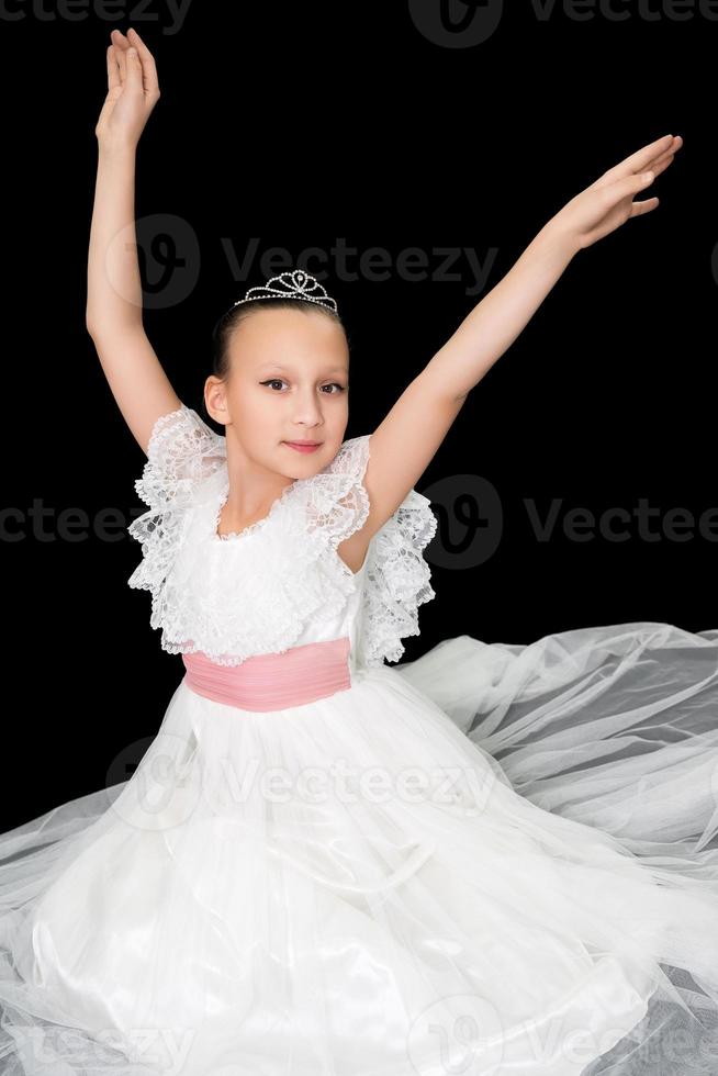 meisje ballerina in wit lang jurk zittend Aan zwart achtergrond en poses elegant met armen verheven foto