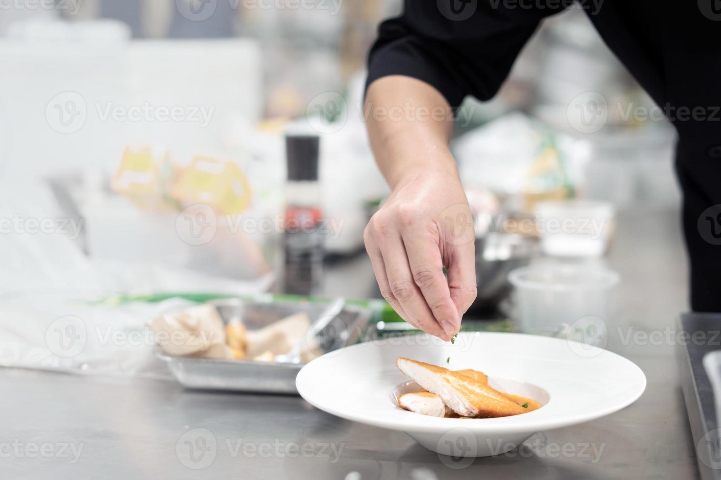 gefocust hand- mannetje chef in zwart uniform versieren voedsel, bestrooi garnituren Aan Koken gebakken steak vis filet , wit tilapia in wit bord in keuken. foto