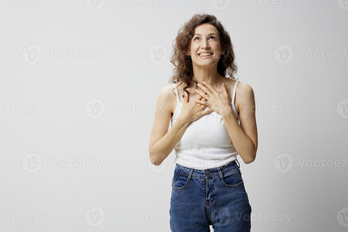 dolblij geschokt verrast gelukkig gekruld mooi vrouw in eenvoudig wit t-shirt vouwen handen Aan borst poseren geïsoleerd Aan over- wit achtergrond. mensen emoties levensstijl concept. kopiëren ruimte foto