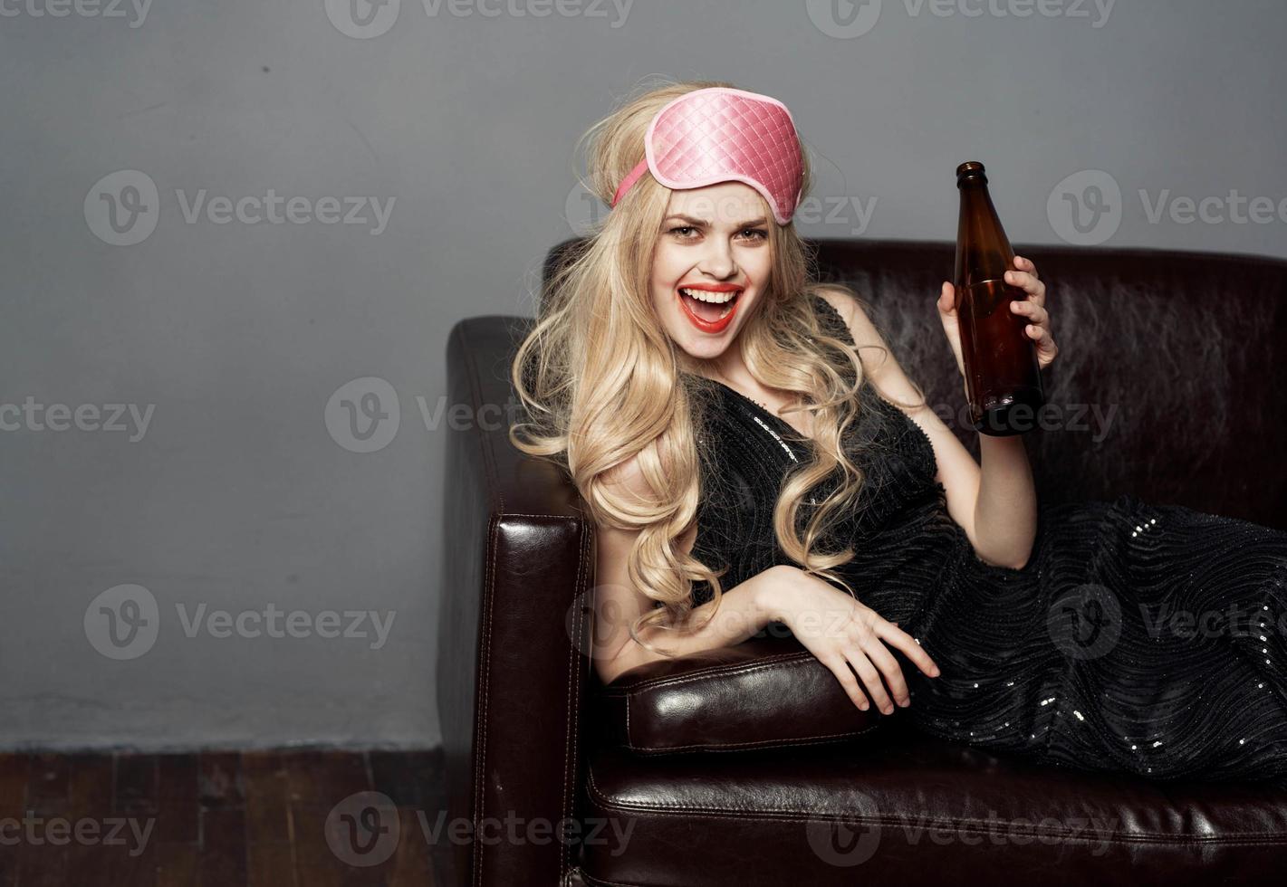 een mooi blond met een fles van bier leugens Aan een leer sofa binnenshuis en een roze masker voor slapen foto