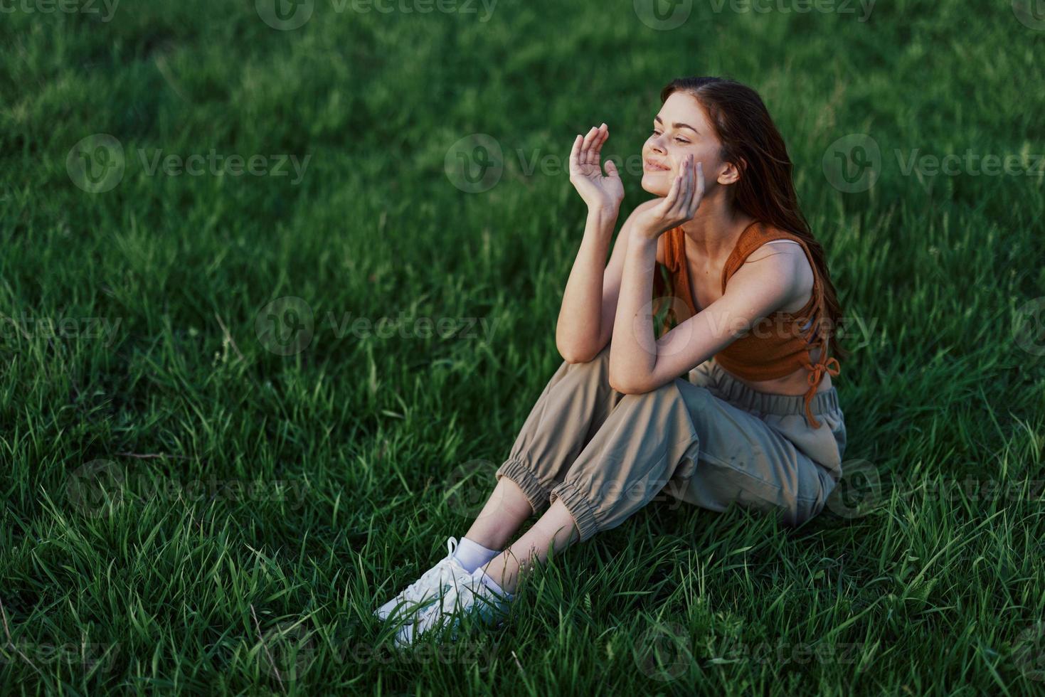 een mooi meisje ontspannende in natuur zittend in broek en top Aan de groen gras in de stralen van de instelling zomer zon. de concept van Gezondheid en zorg voor de lichaam en de milieu foto